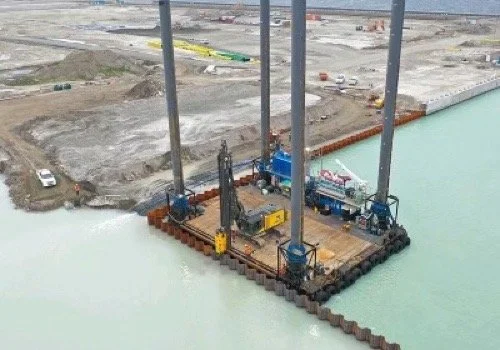 Construction site on water with large machinery and tall poles, surrounded by a barrier.