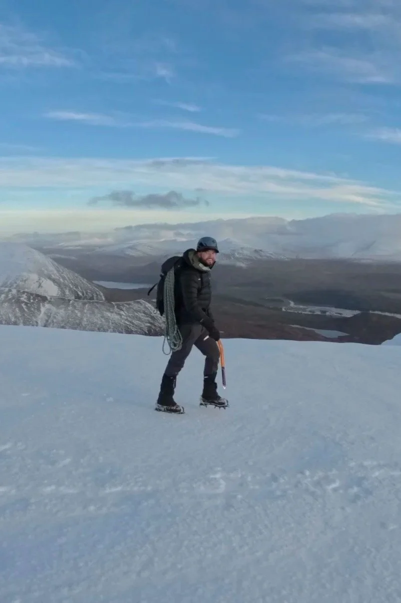 A man in winter gear, including a helmet and backpack, climbing snow-covered terrain with an ice axe, surrounded by a snowy mountainous landscape and cloudy sky.