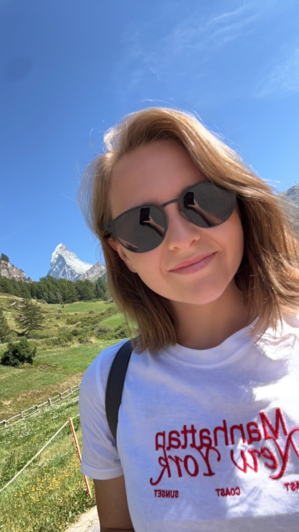 A woman with shoulder-length brown hair and sunglasses standing outdoors with green fields, trees, and mountains in the background.
