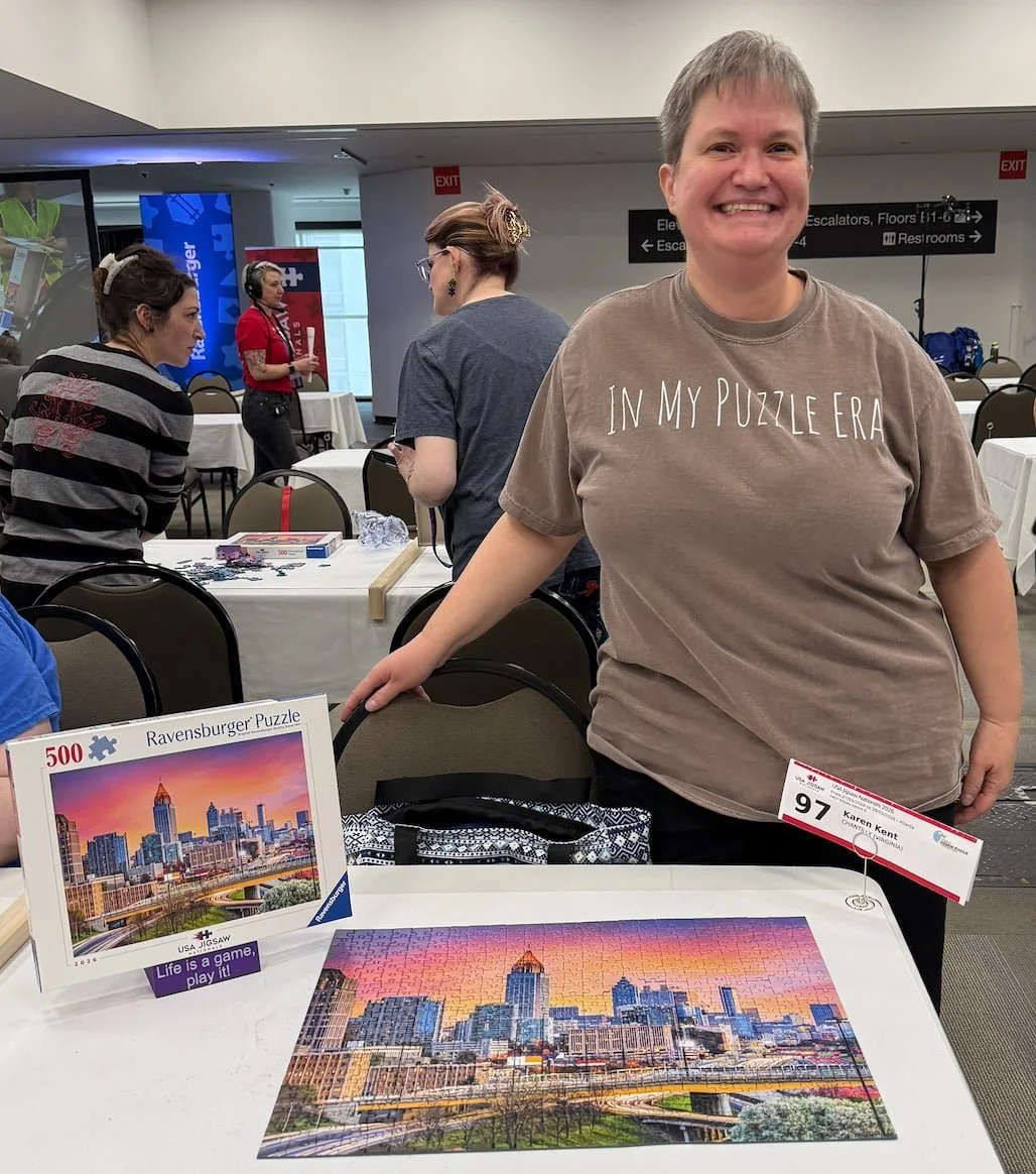 A woman smiling at a table with a completed 500-piece Ravensburger puzzle of a city skyline. Other people are in the background at the puzzle event in a conference room.