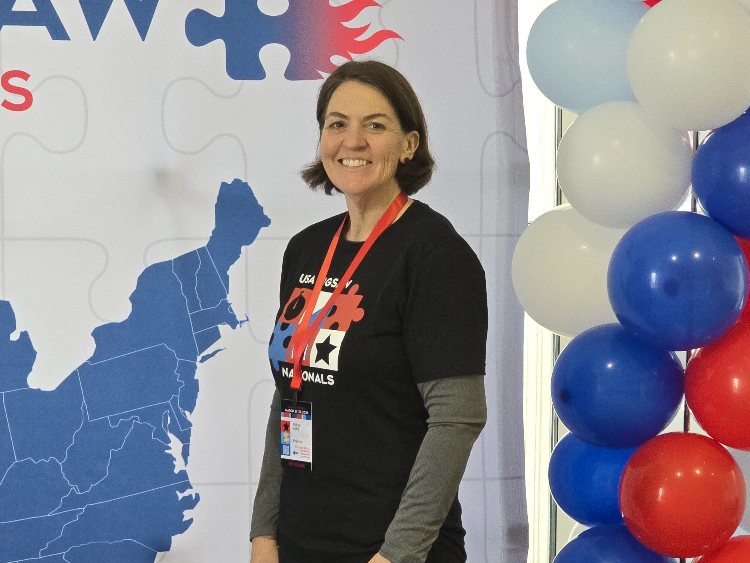 A woman smiling at an event with a USA-themed background, balloons in red, white, and blue, and wearing a black t-shirt with a puzzle piece and star design.