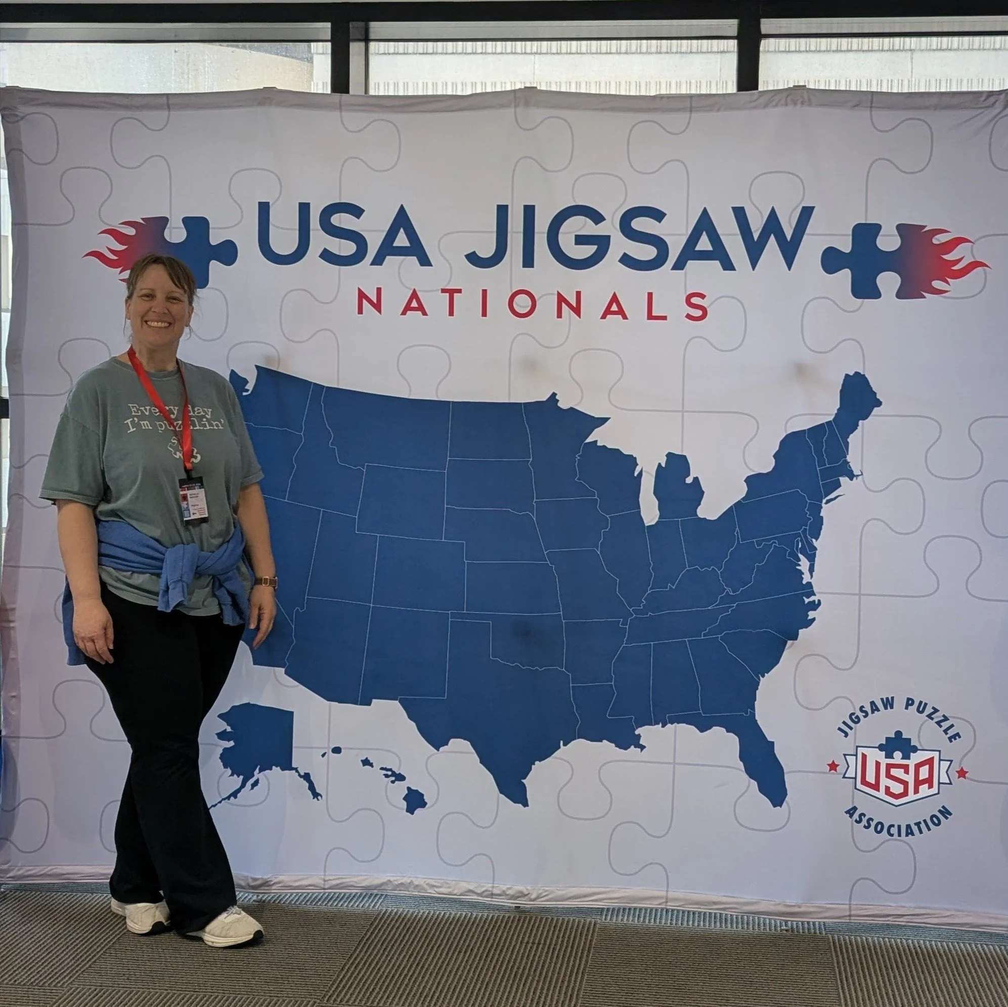 A woman standing in front of a large USA map made of puzzle pieces at a jigsaw puzzle event. The sign reads 'USA Jigsaw Nationals' with a logo that says 'Jigsaw Puzzle USA Association.' The woman is smiling, wearing a gray T-shirt, black pants, and white shoes, with a red lanyard around her neck.