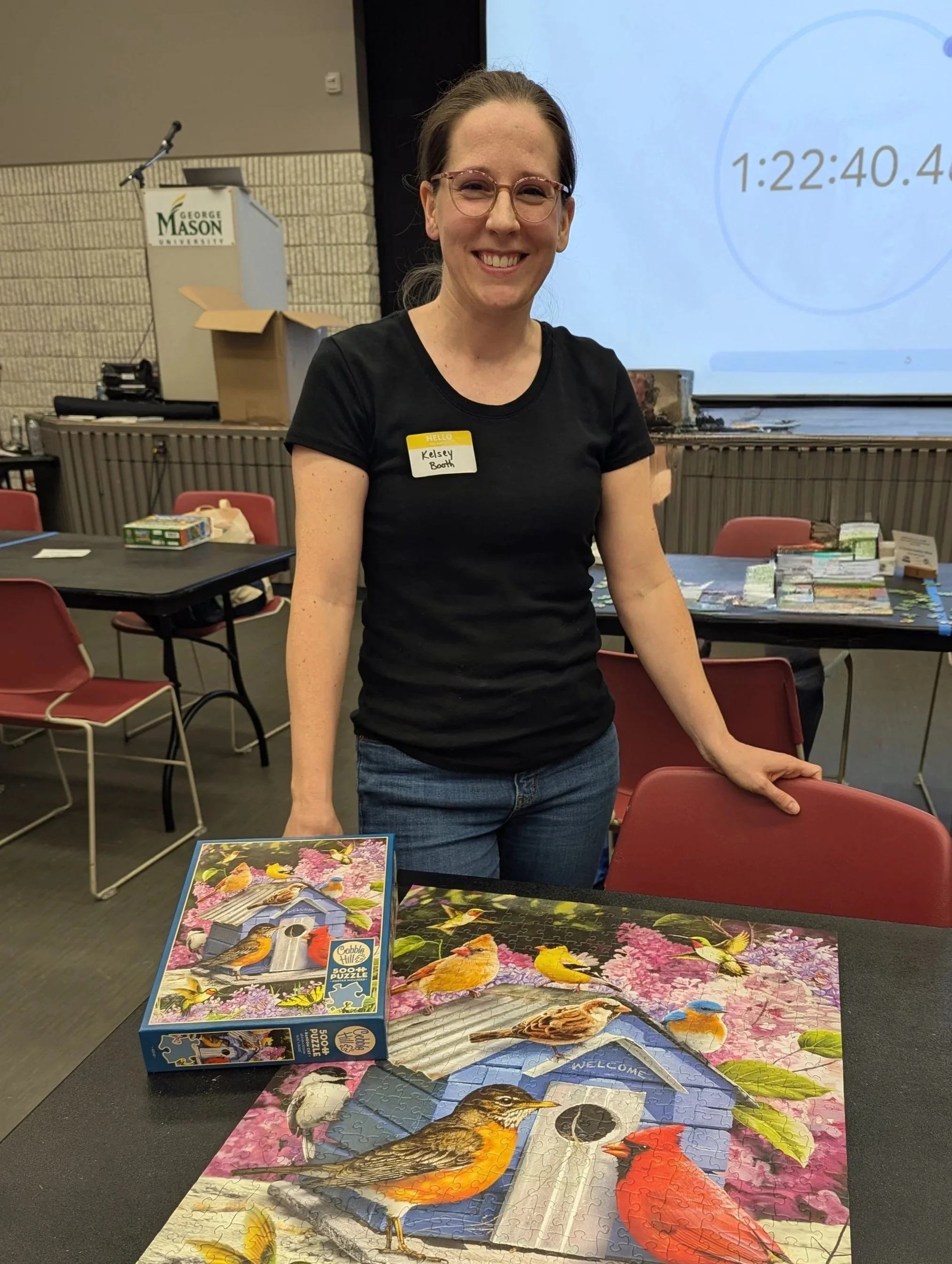 A woman with glasses and a black shirt smiling at a table with a completed birdhouse puzzle and a box of birdhouse puzzle pieces. She has a yellow name badge that reads 'Kelsey Booth'. The background shows tables, chairs, and a stage area with a large screen.