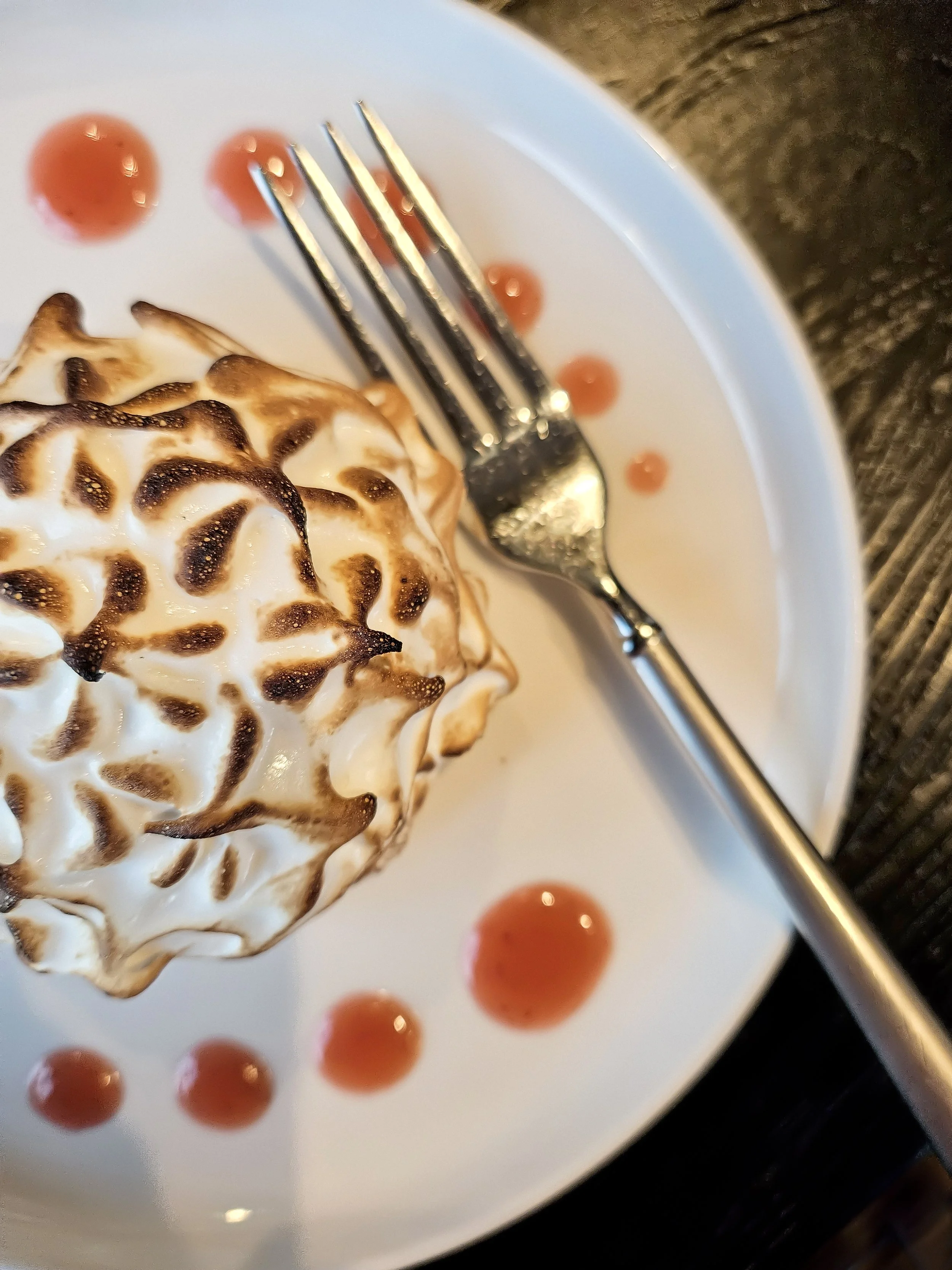 A close-up of a toasted meringue dessert on a white plate, with red sauce drizzles and a silver fork resting on the plate, placed on a dark wooden surface.