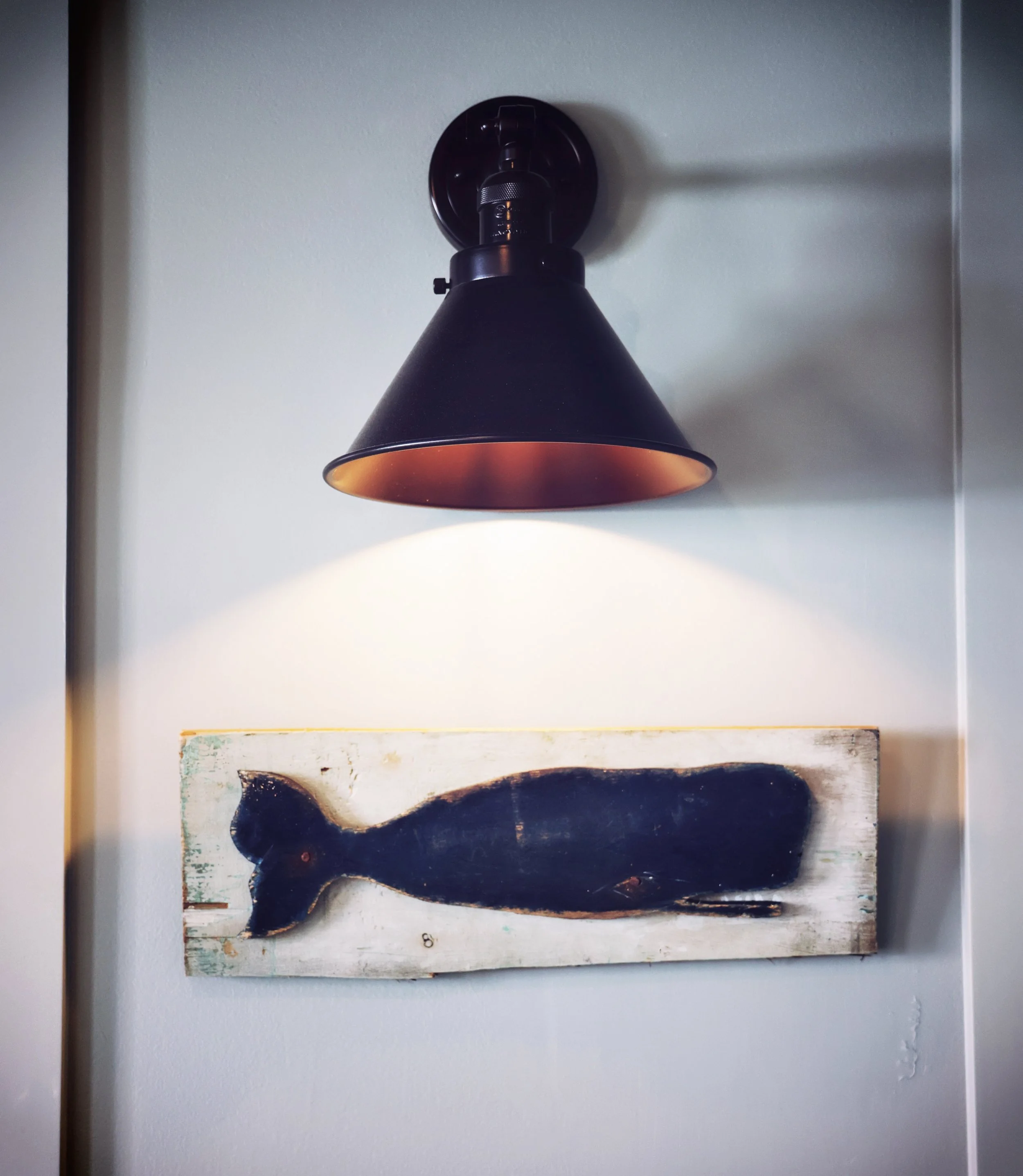 Wall-mounted black lamp shining light onto a wooden fish-shaped decoration on a white wall.