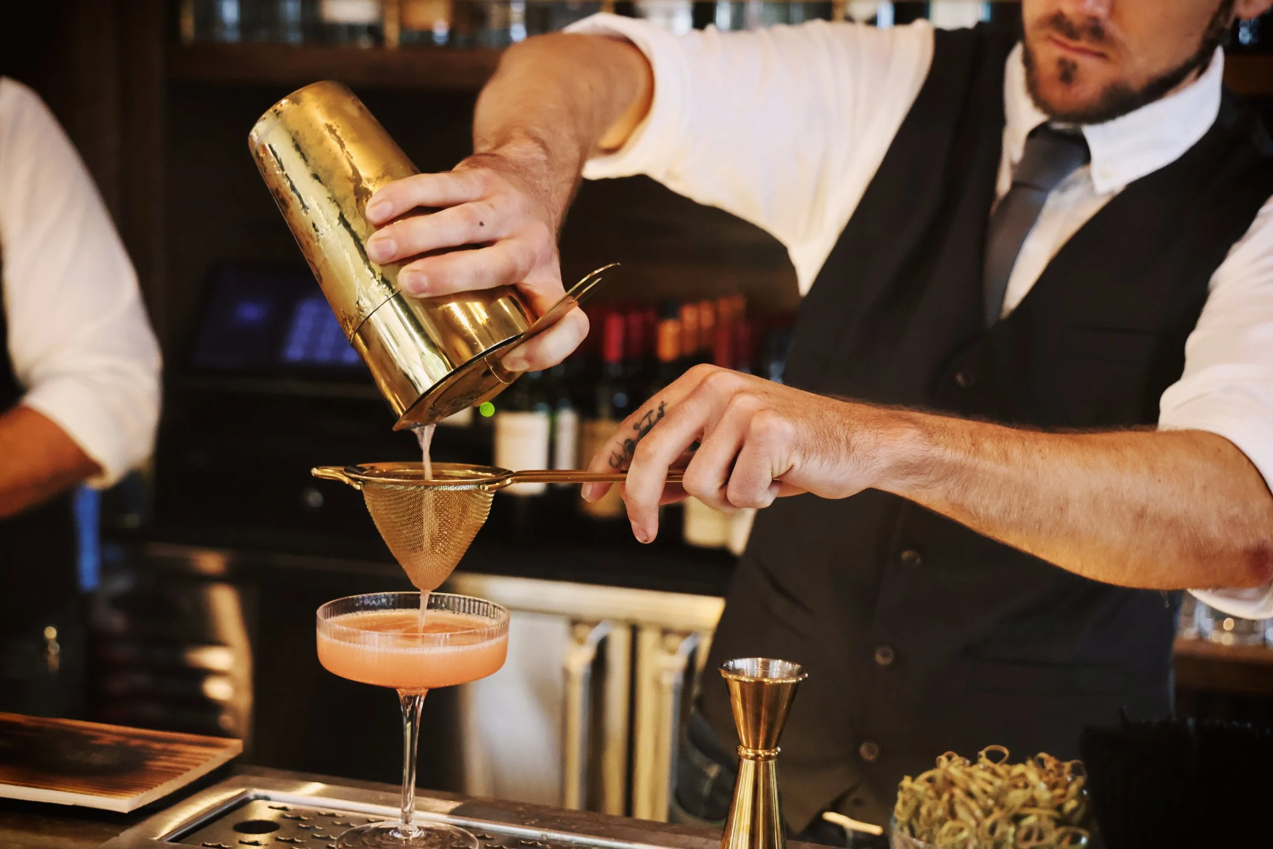 A bartender pouring liquid through a fine mesh strainer into a pink cocktail in a coupe glass at Jeremiah's Restaurant.