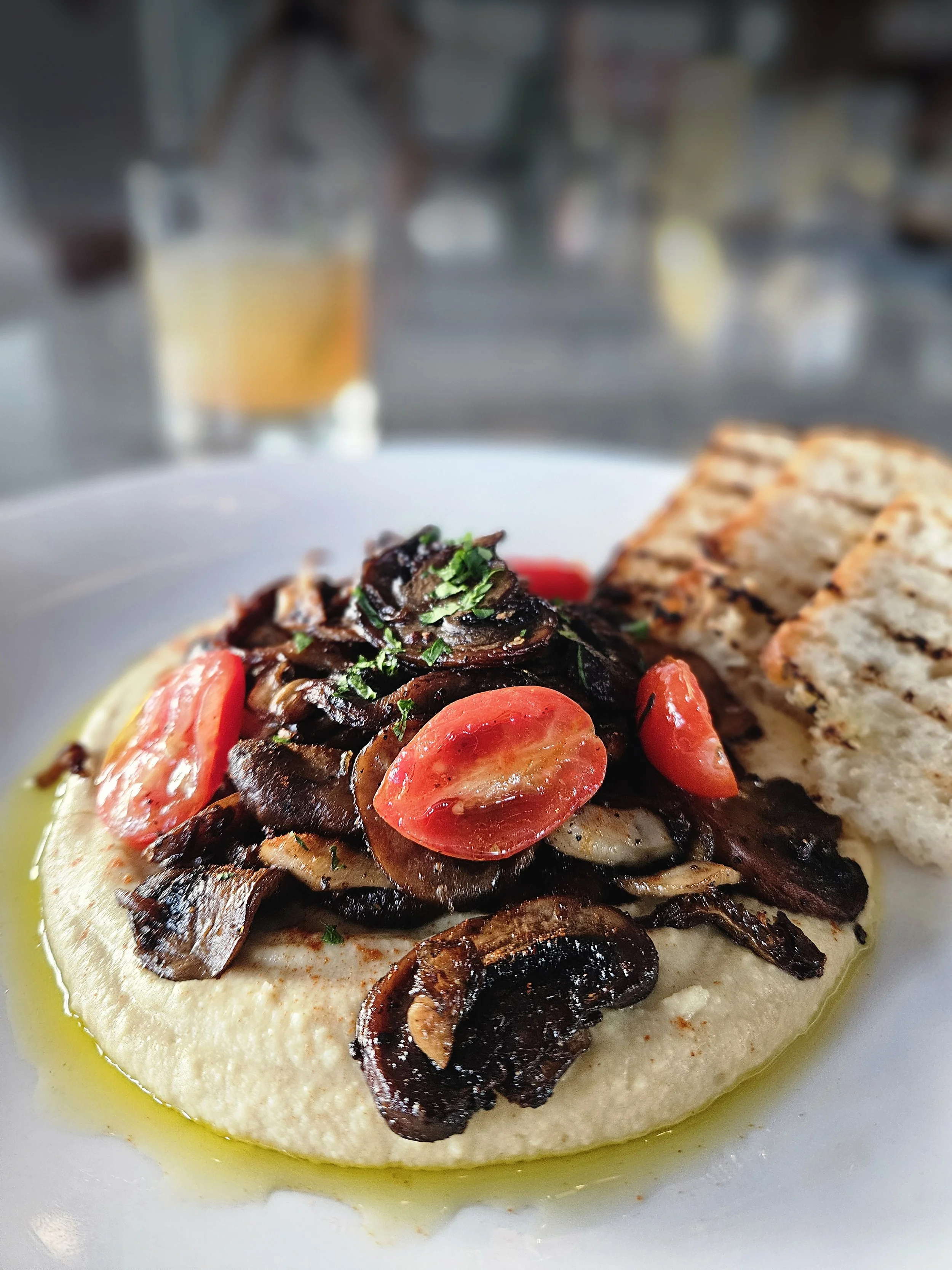 Dish with sautéed mushrooms, cherry tomatoes, hummus, and grilled bread on a white plate.