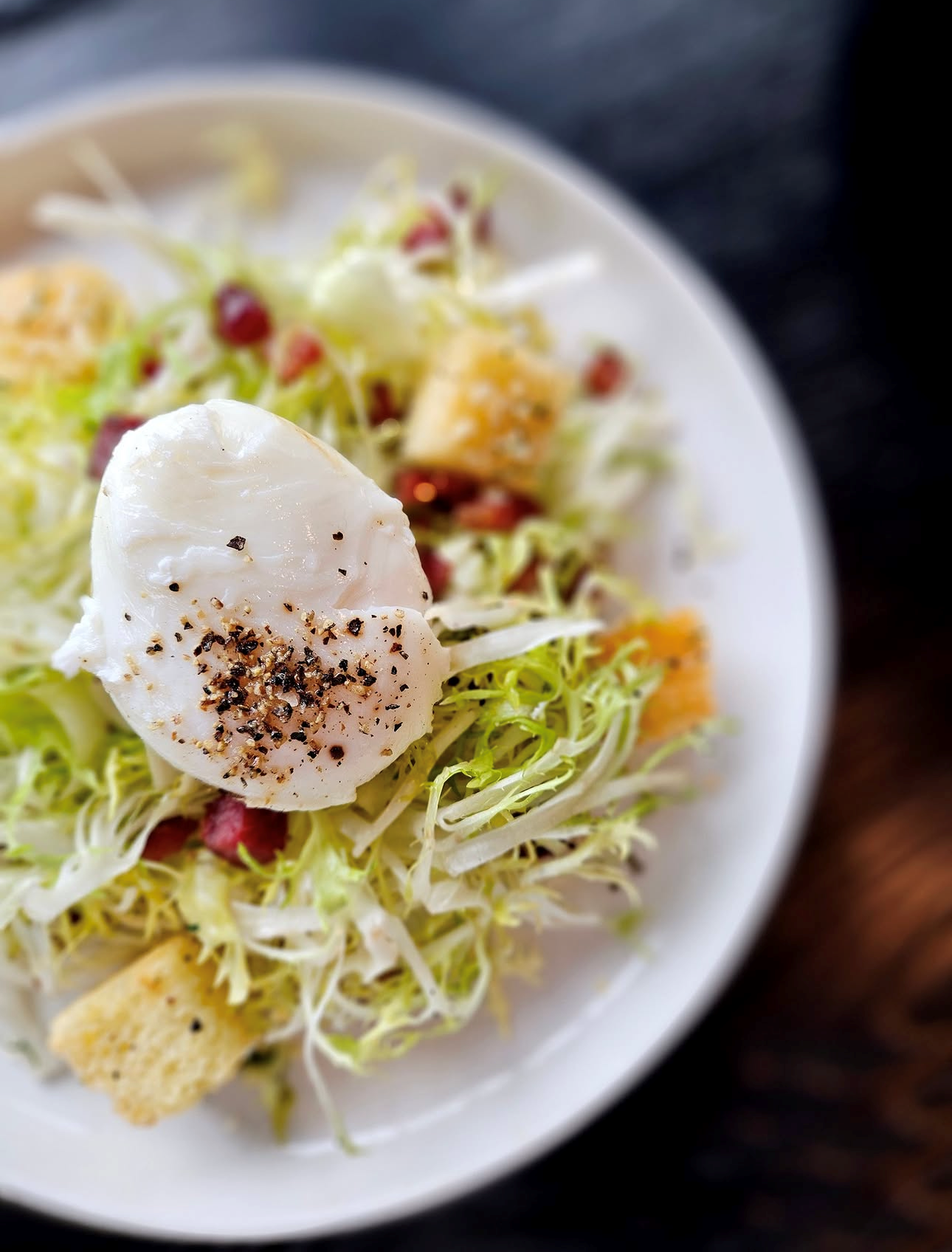Close-up of a salad with shredded lettuce, croutons, bacon bits, and a poached egg seasoned with black pepper on top.