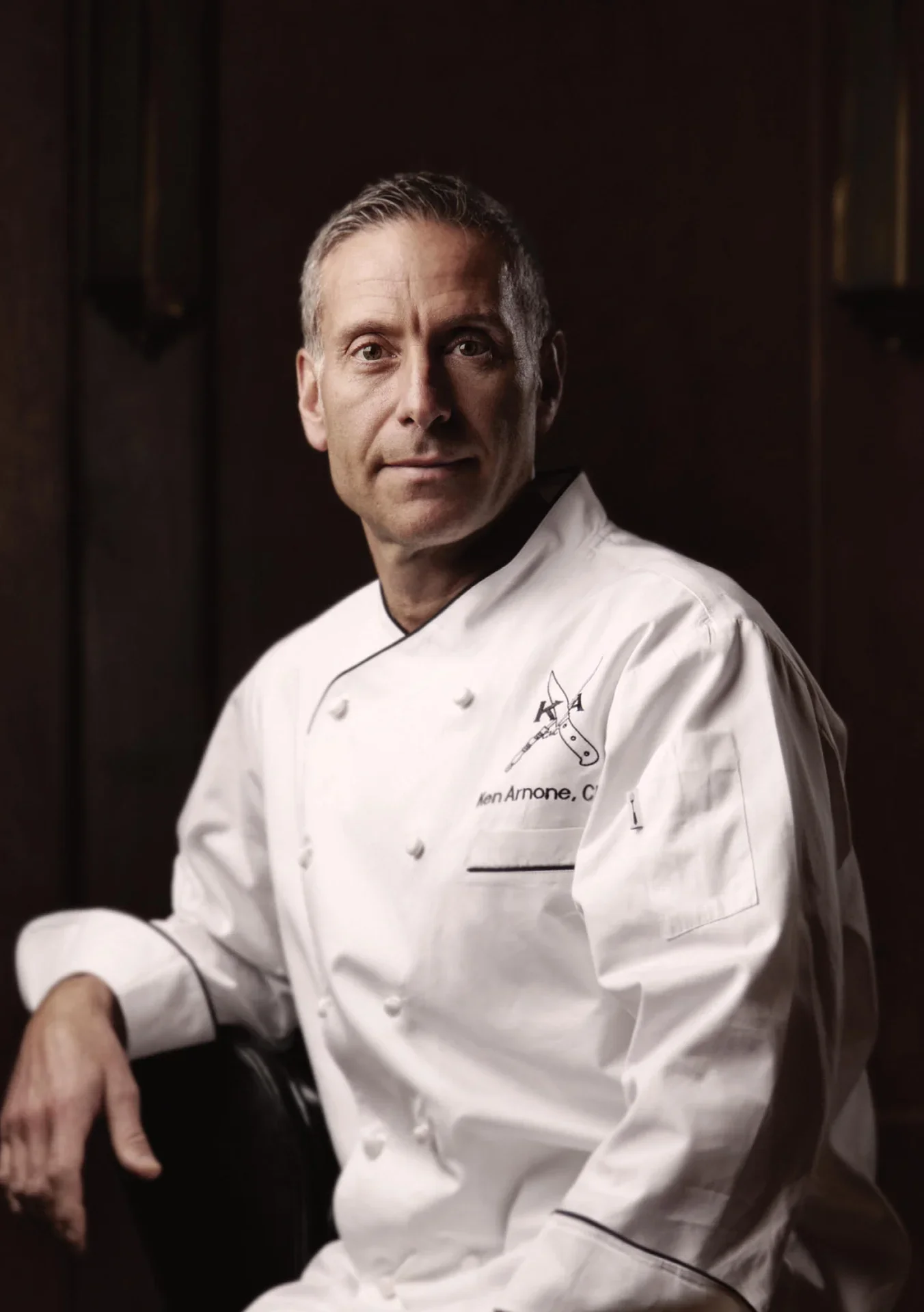 Chef Ken Arnone, Certified Master Chef and Cannonball Hospitality Corporate Chef, in a white chef's coat with embroidery, sitting in a dark room, looking at the camera.