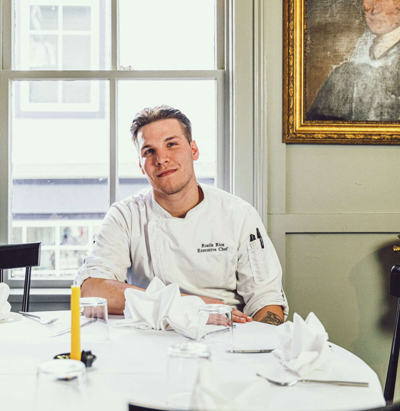 Chef Roelle Rios, head chef of Jeremiah's Restaurant in Mystic, sitting at a table in a restaurant, wearing a white chef's coat with his name and title embroidered on it, with a window behind him and a portrait on the wall to his right.