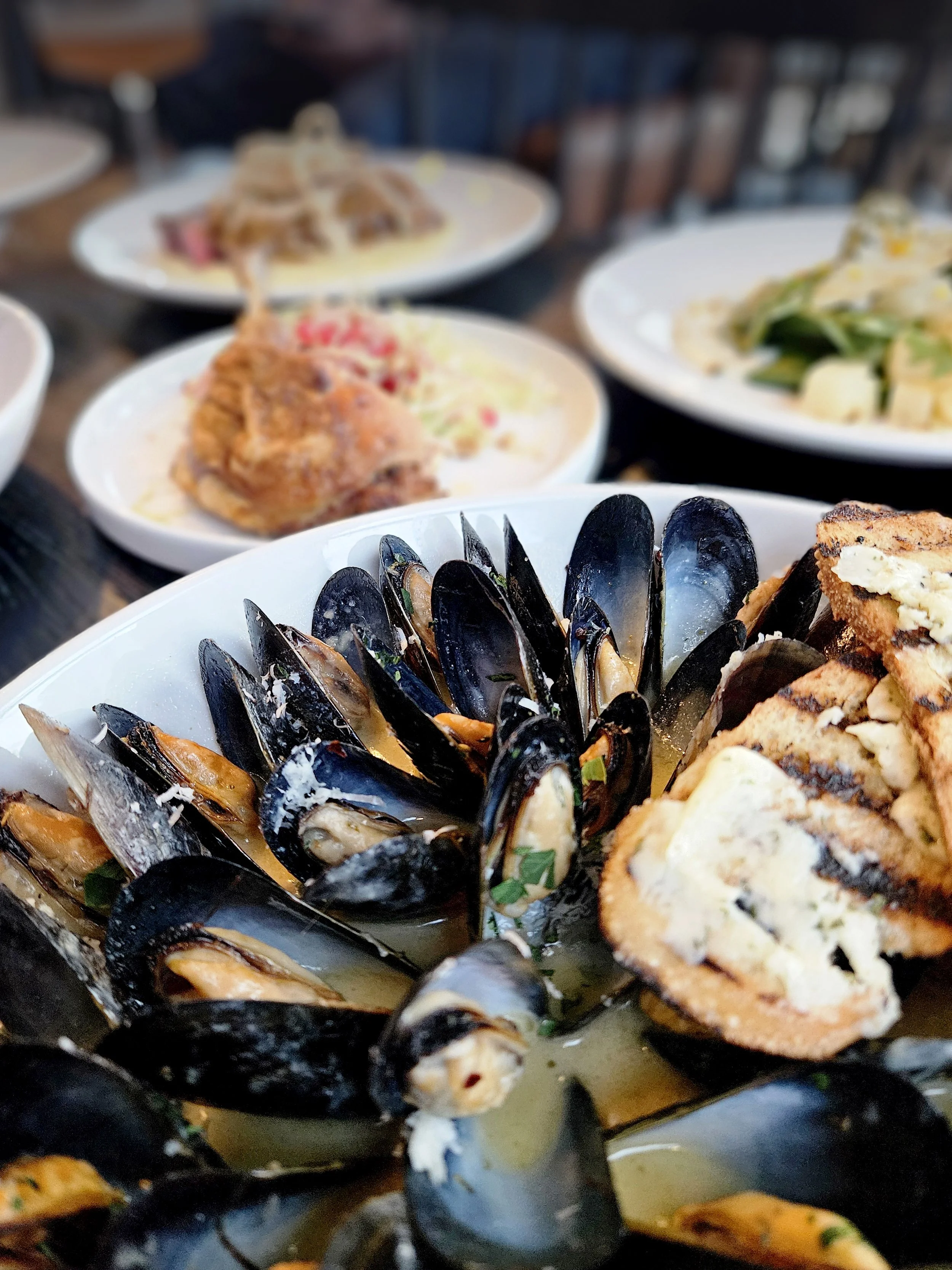 A bowl of cooked mussels with herbs and broth, with slices of bread or toast nearby, surrounded by various other dishes on a wooden table.