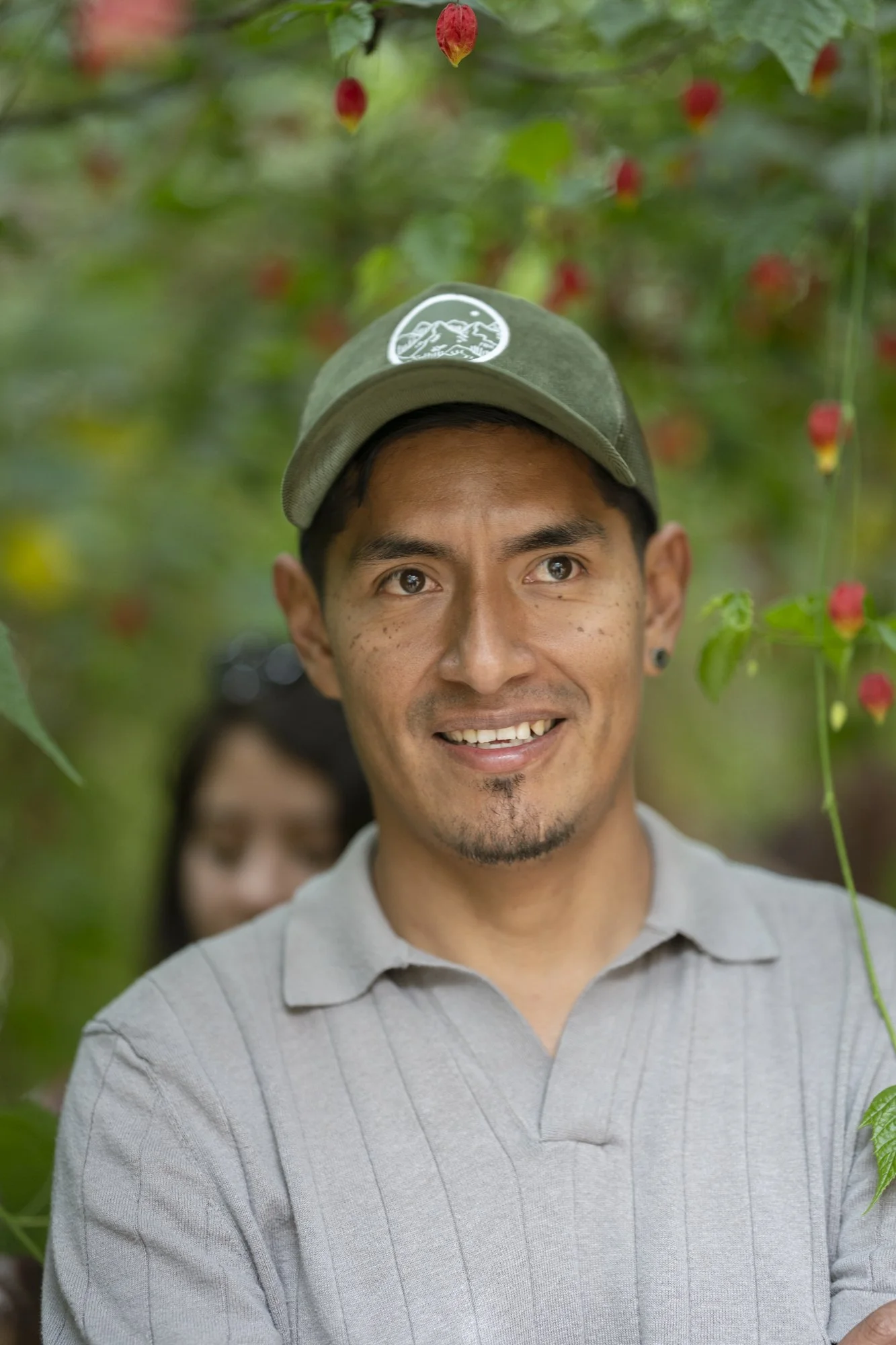 Hombre sonriendo en un entorno de plantas con flores rojas colgantes.