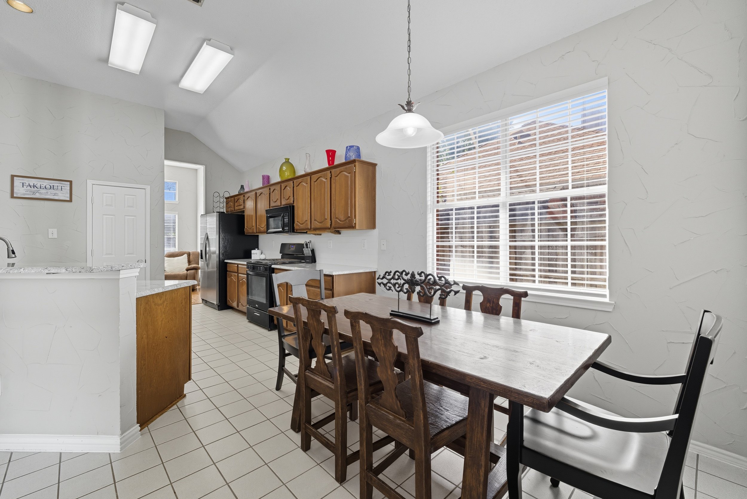 Kitchen with wooden cabinets, a dining table with chairs, large window with blinds, and a hanging light fixture.