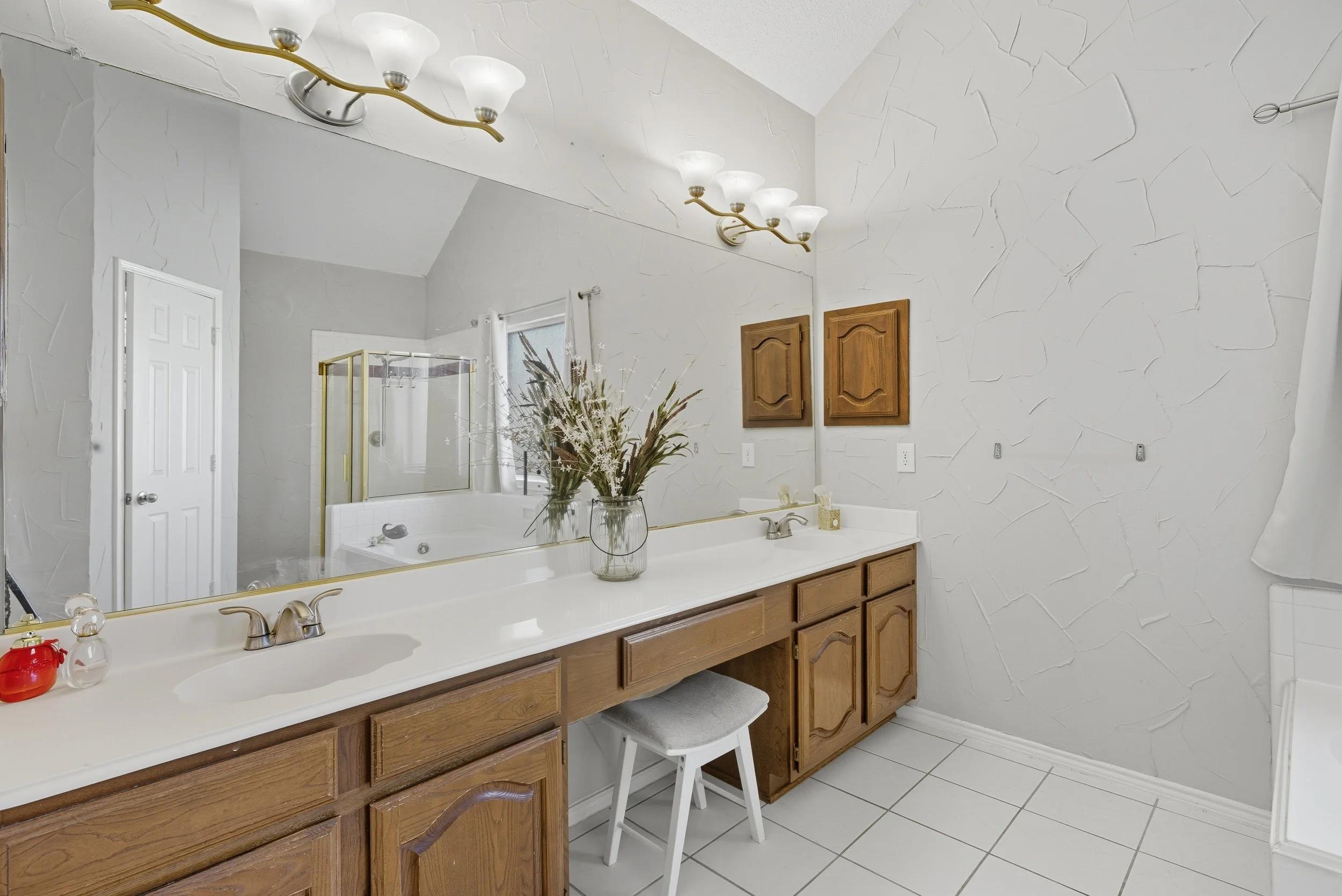 Bathroom with a large mirror, wooden cabinets, a double sink vanity, a vase with dried flowers, wall-mounted lights, a shower, and a bathtub with a window.