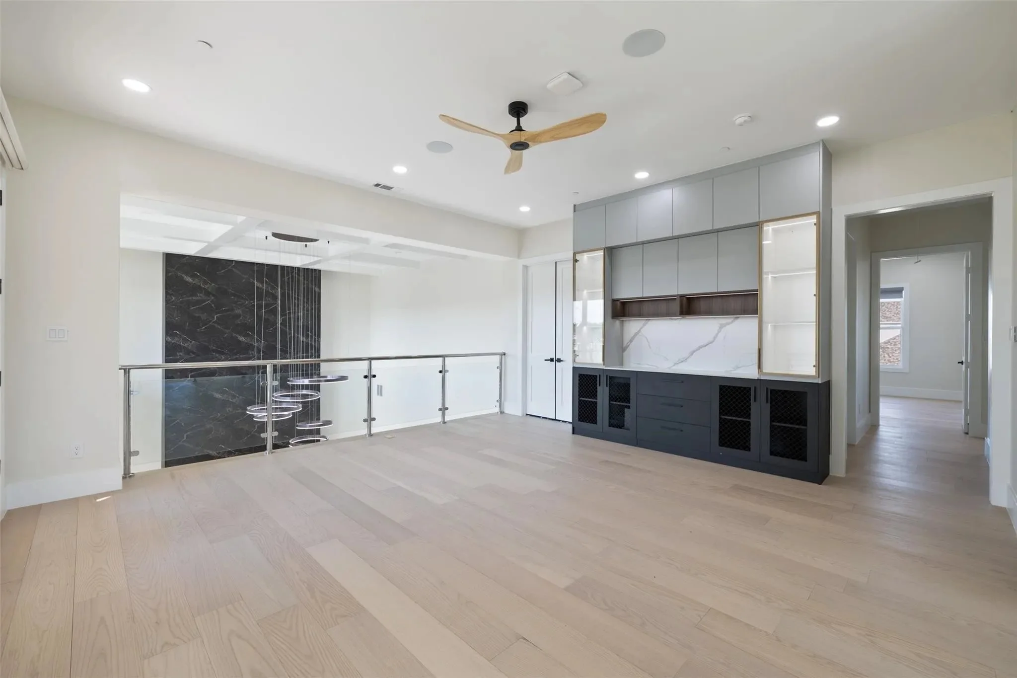 Bright, modern living room with light wood flooring, built-in gray and black cabinetry, glass shelving, white marble backsplash, ceiling fan with wooden blades, and recessed lighting.