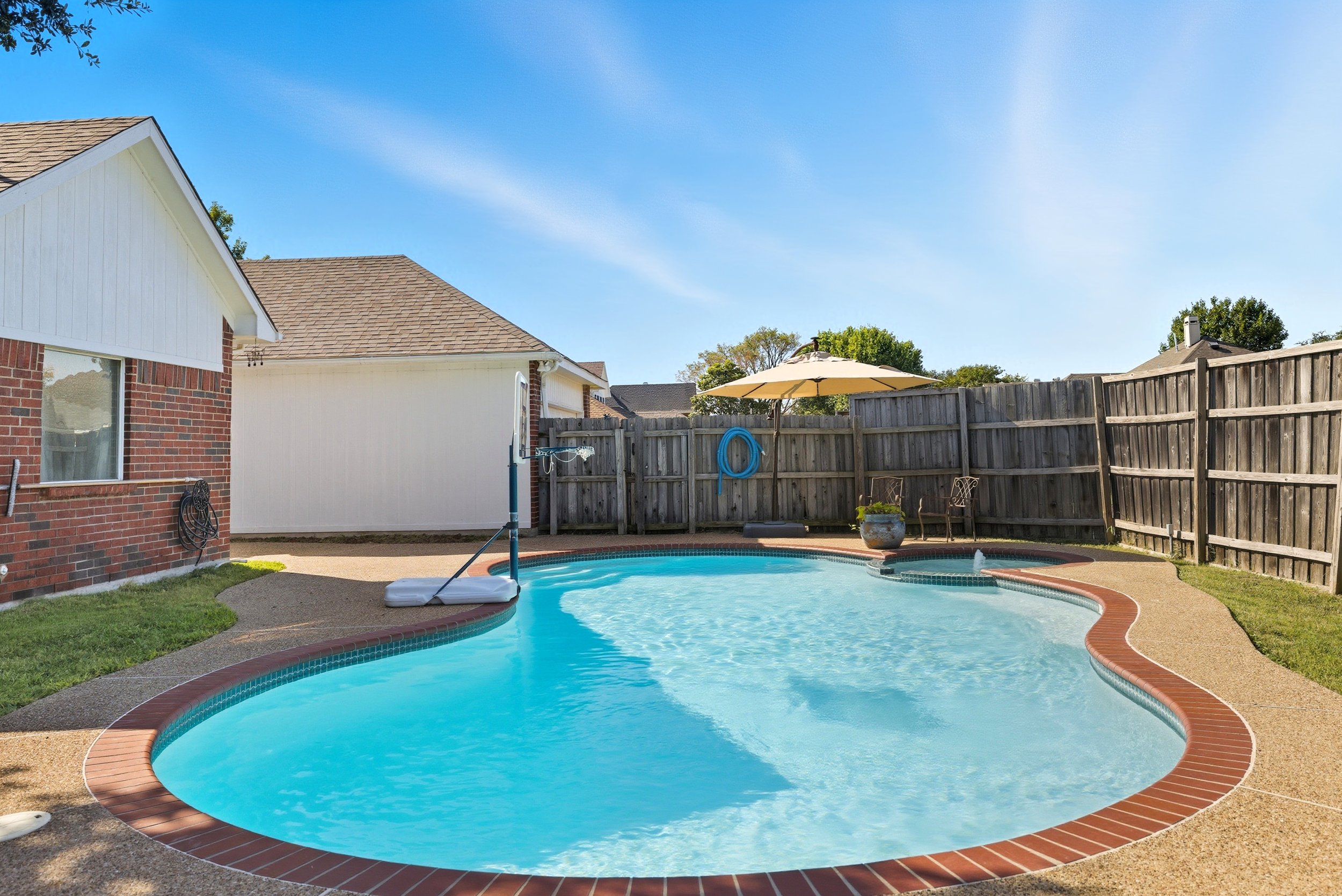 A backyard with a freeform swimming pool, a wooden privacy fence, a patio umbrella, and outdoor chairs under a blue sky.