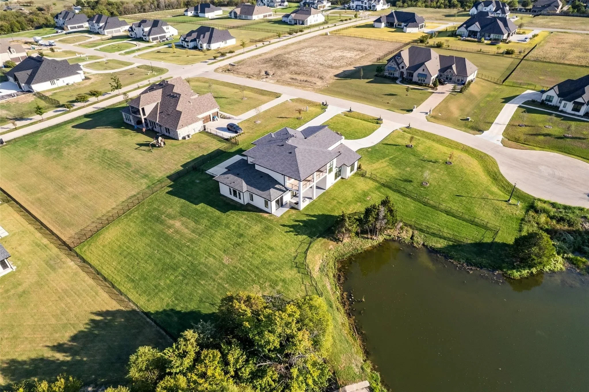 Aerial view of a suburban neighborhood with houses, green lawns, paved roads, and a small pond, highlighting a white two-story house with a gray roof and fenced backyard.