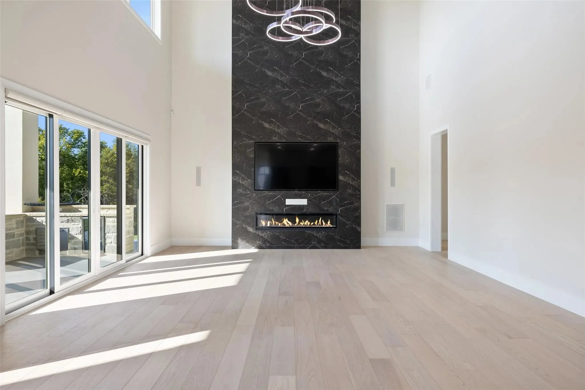 Modern living room with a black marble accent wall, mounted TV, and a built-in fireplace, with large sliding glass doors letting in sunlight onto light-colored hardwood floors.