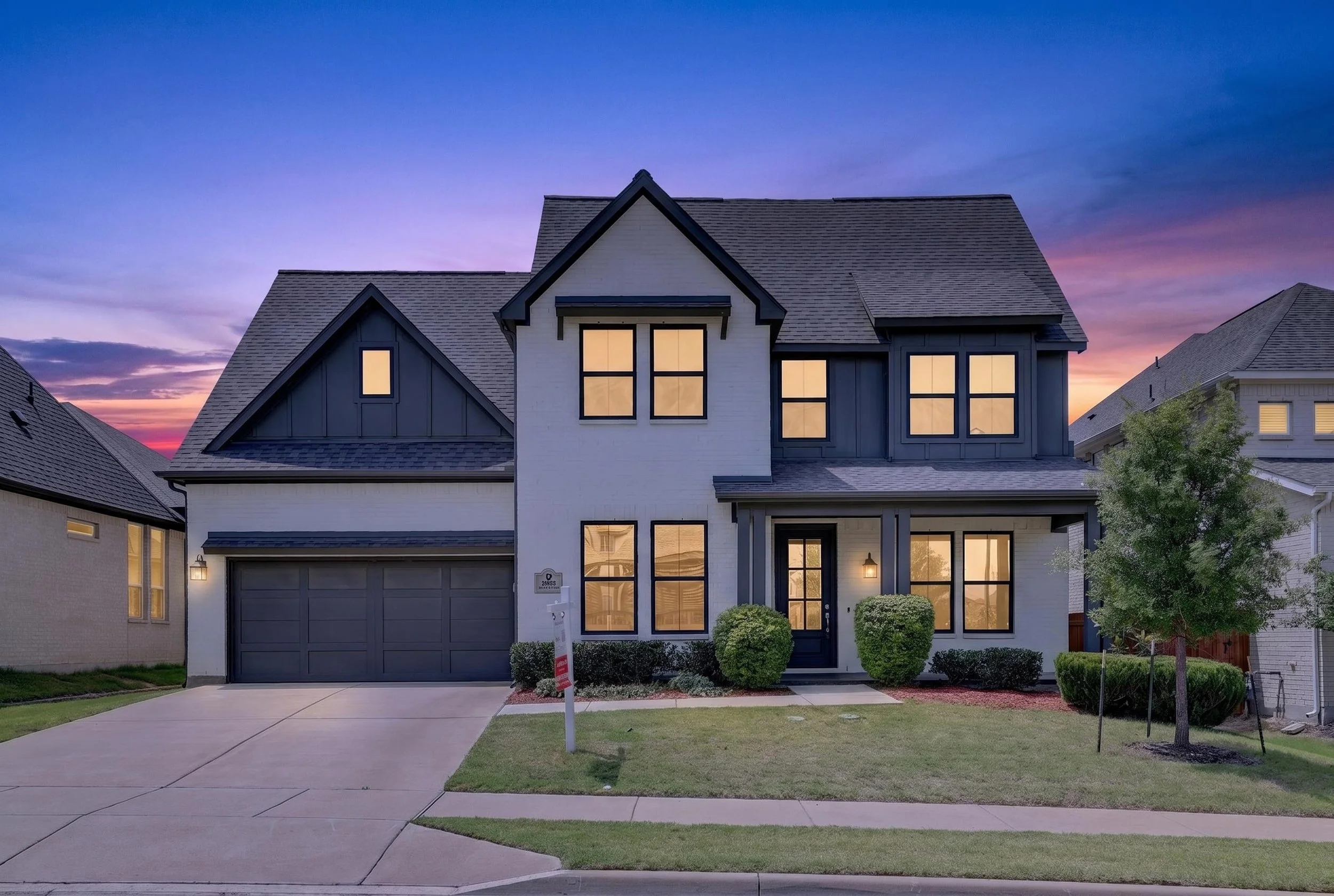 Modern two-story house with a gray garage door, large windows, and a front porch, illuminated by warm exterior lights during dusk. Neighborhood houses visible on both sides, manicured lawn, and a sidewalk.