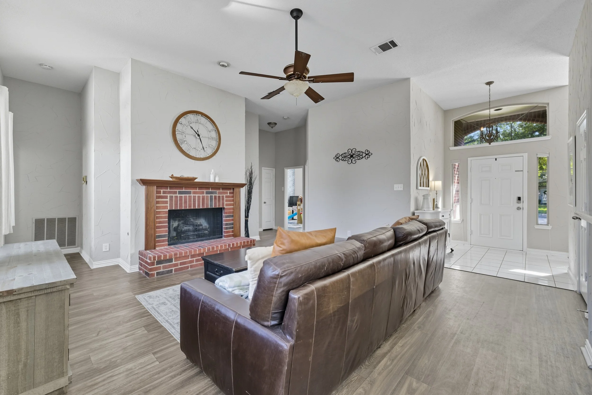 Living room with brown leather sofa, brick fireplace with a large clock above, ceiling fan, white walls, and entrance area with tile flooring and outdoor view.