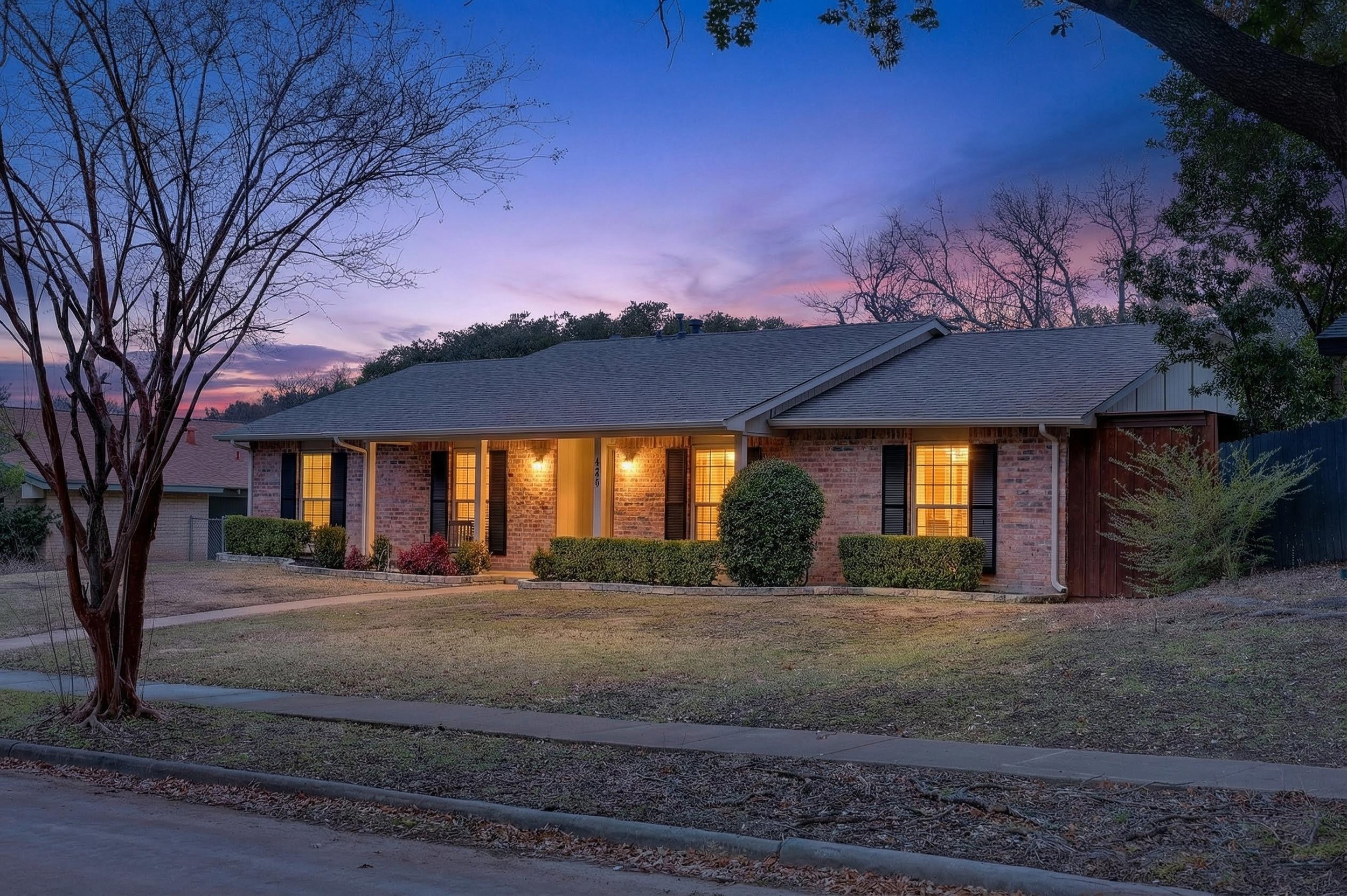 A single-story brick house with a dark roof illuminated by warm exterior lights, surrounded by a well-maintained lawn and trimmed bushes, during sunset with a colorful sky and leafless trees in the background.