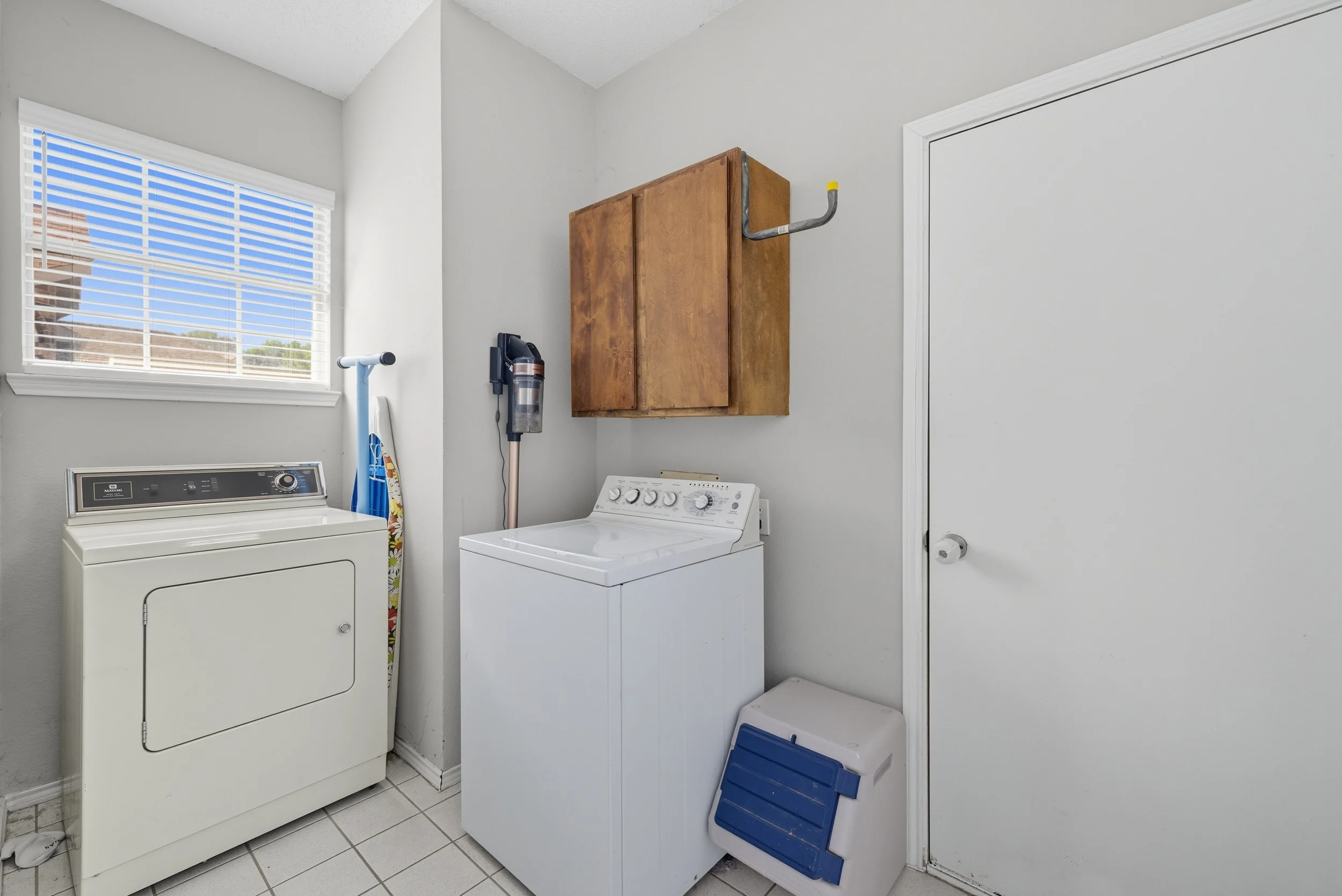 Laundry room with a top-loading washing machine, a dryer, a wall-mounted wooden cabinet, an ironing board, a cordless vacuum, and a small pet door.