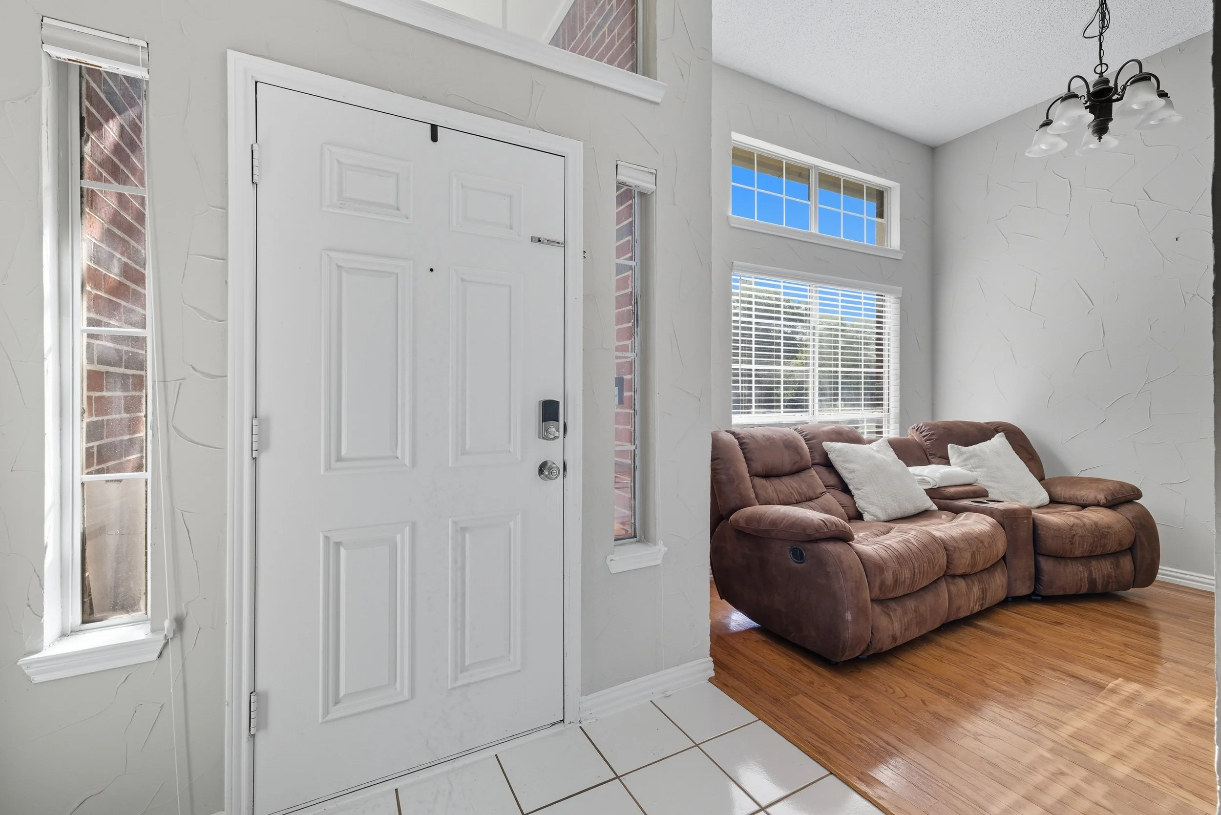 Interior of a house showing an entrance door with a keypad lock, a window with blinds, a brown sofa with pillows, and a wooden floor in a bright room.