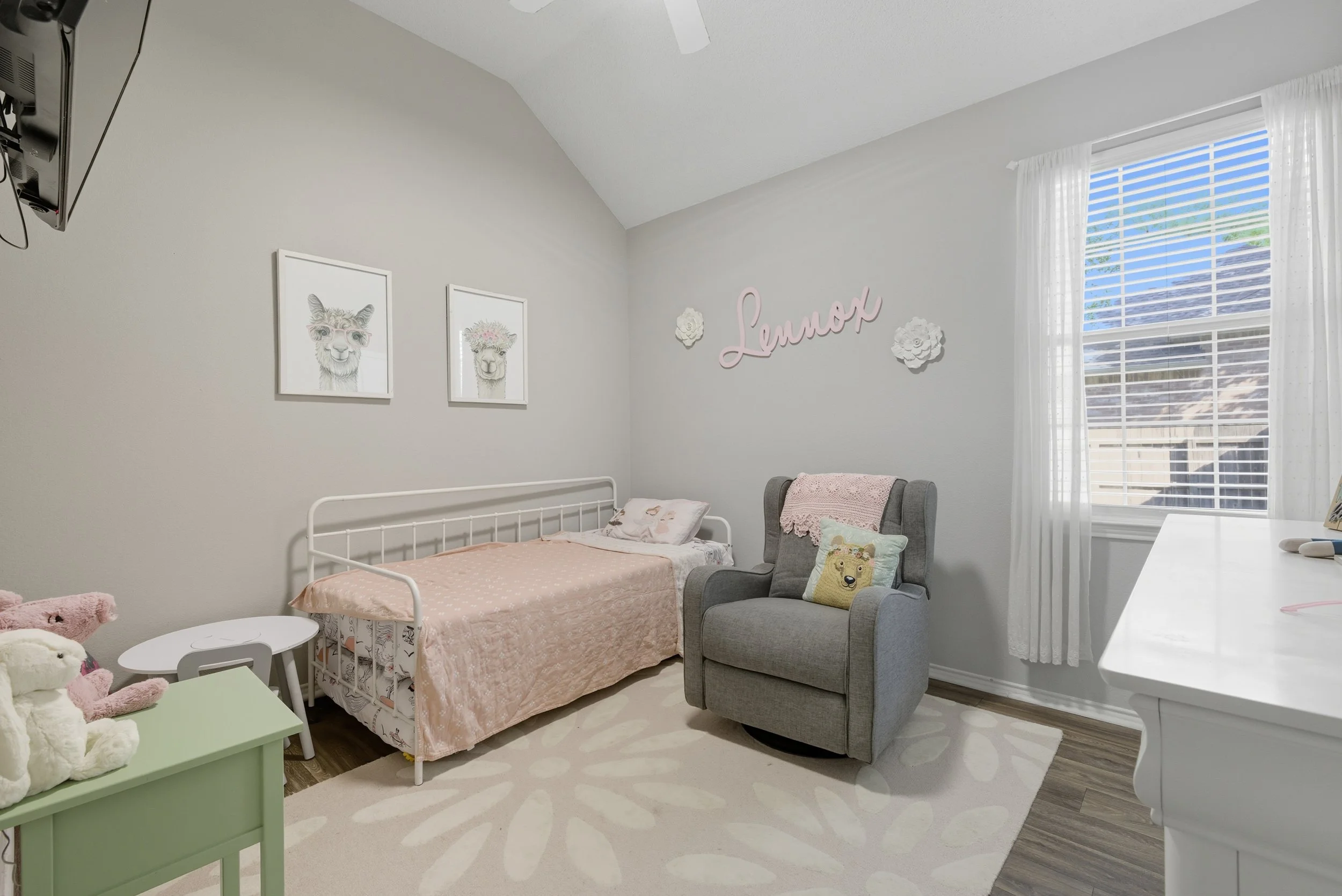 A cozy children's bedroom featuring a white bed with pink bedding, a gray armchair with a bear pillow, and framed animal illustrations on the wall, with a window dressed in white curtains letting in natural light and a decorative pink sign that says 