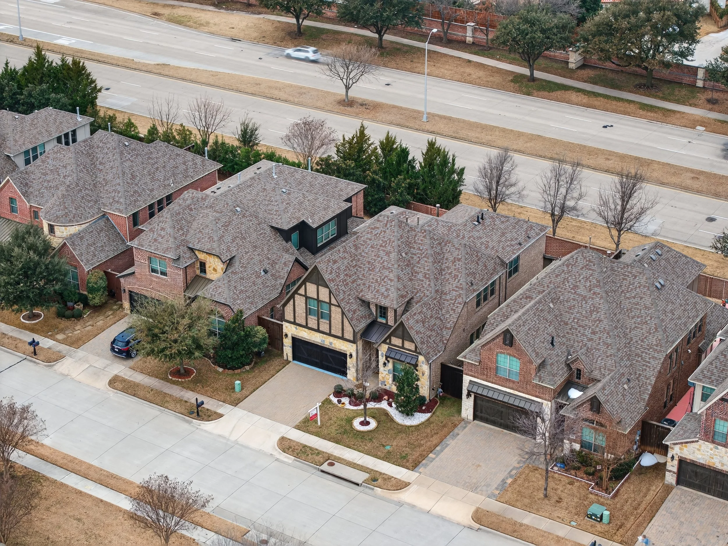 Aerial view of a suburban neighborhood with several detached houses, some with brick exteriors and others with wood siding, featuring tiled roofs. There are streets with parked cars, trees, and manicured lawns.