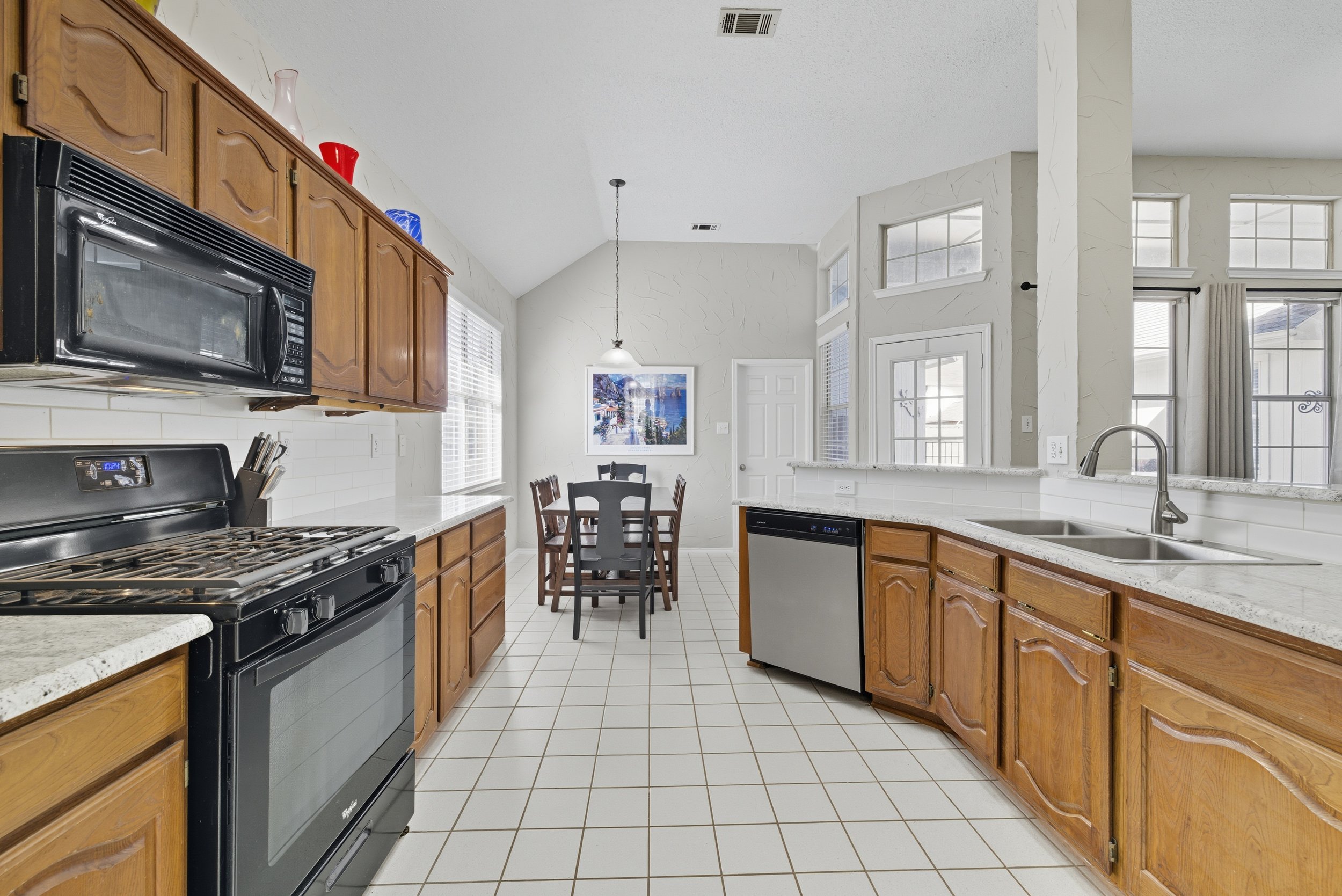 Kitchen with wooden cabinets, white countertops, and a black stove. There is a microwave above the stove, a dishwasher, and a double sink. A small dining table with four chairs is in the background near a window and artwork on the wall. Large windows