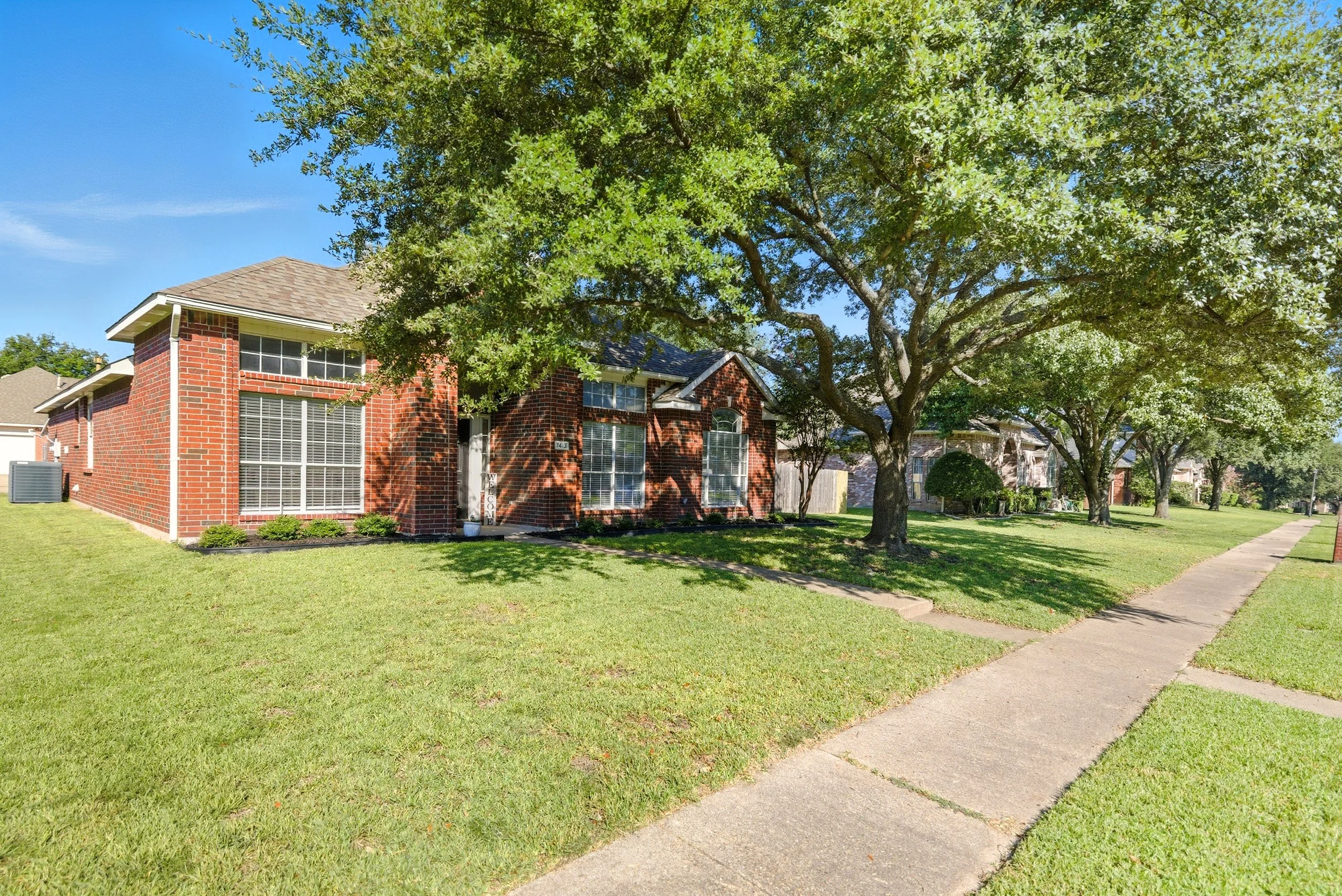 A suburban neighborhood with brick houses, lush green grass, large leafy trees, and a sidewalk on a sunny day.