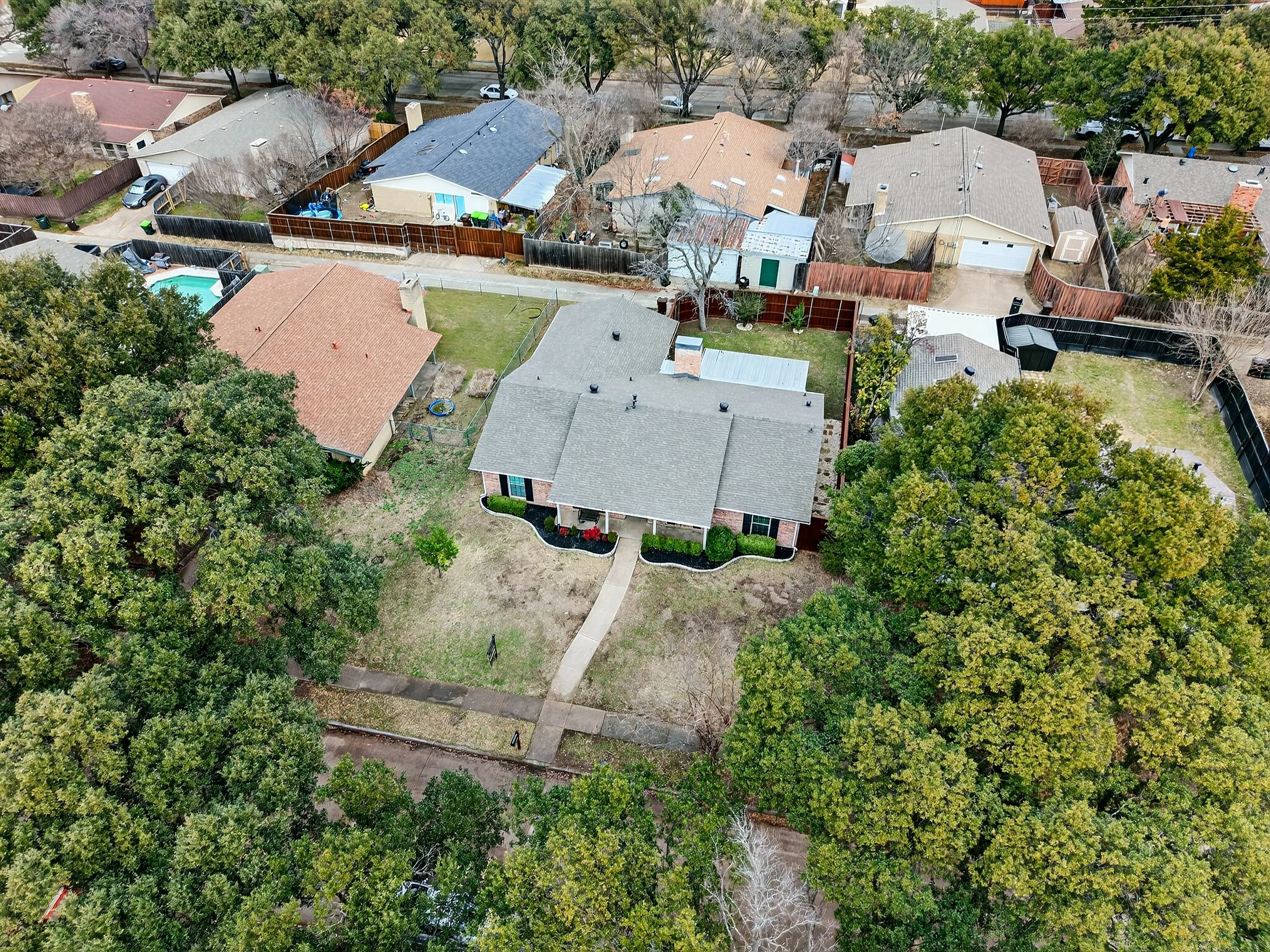An aerial view of a suburban neighborhood showing multiple houses with backyard fences, trees, and a backyard with a pool, lawn, and patio.