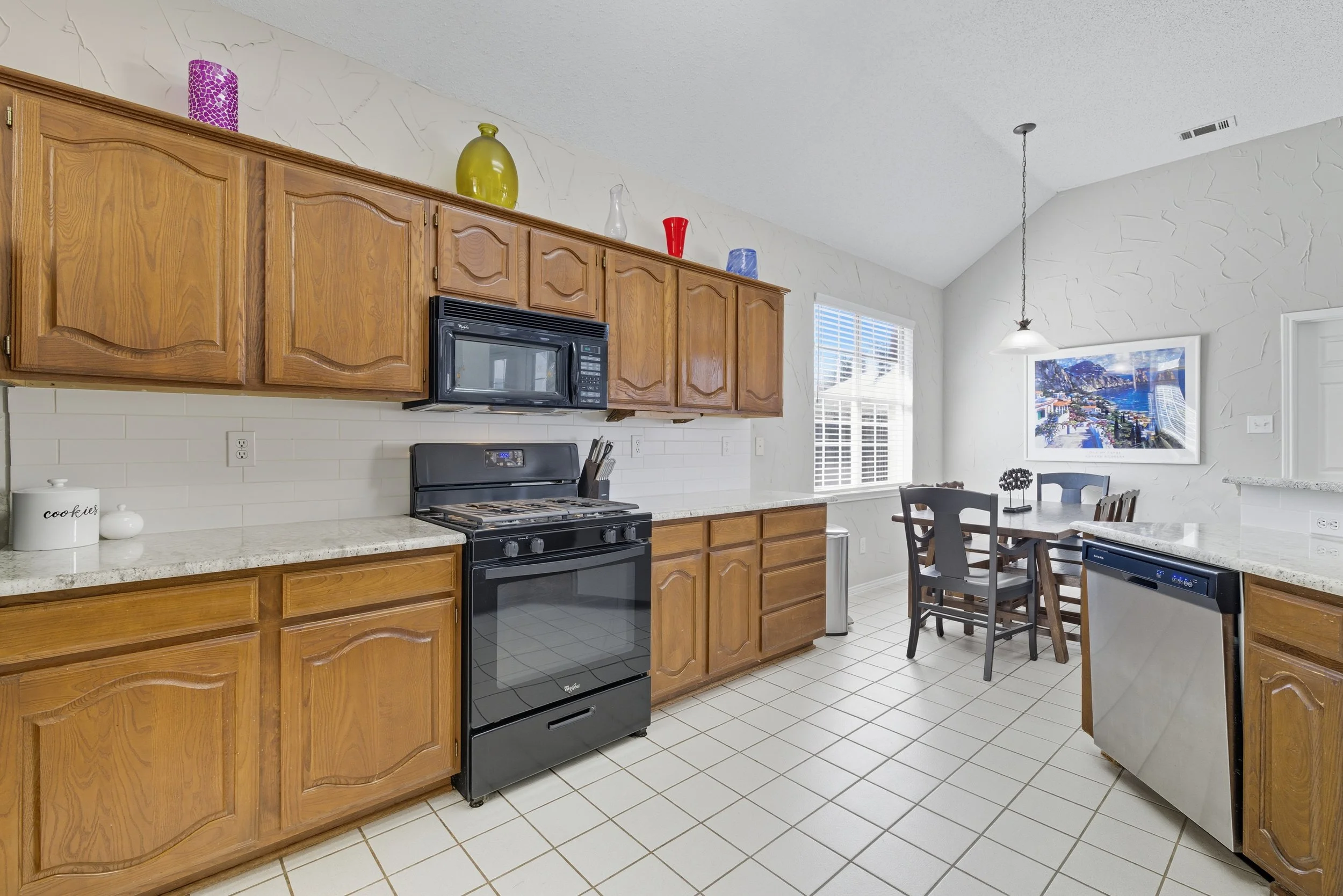 A kitchen with wooden cabinets, black stove, microwave, and dishwasher. There is a dining area with a table and chairs, and a large framed picture on the wall. The kitchen has white tiled floor and walls with a window letting in natural light.