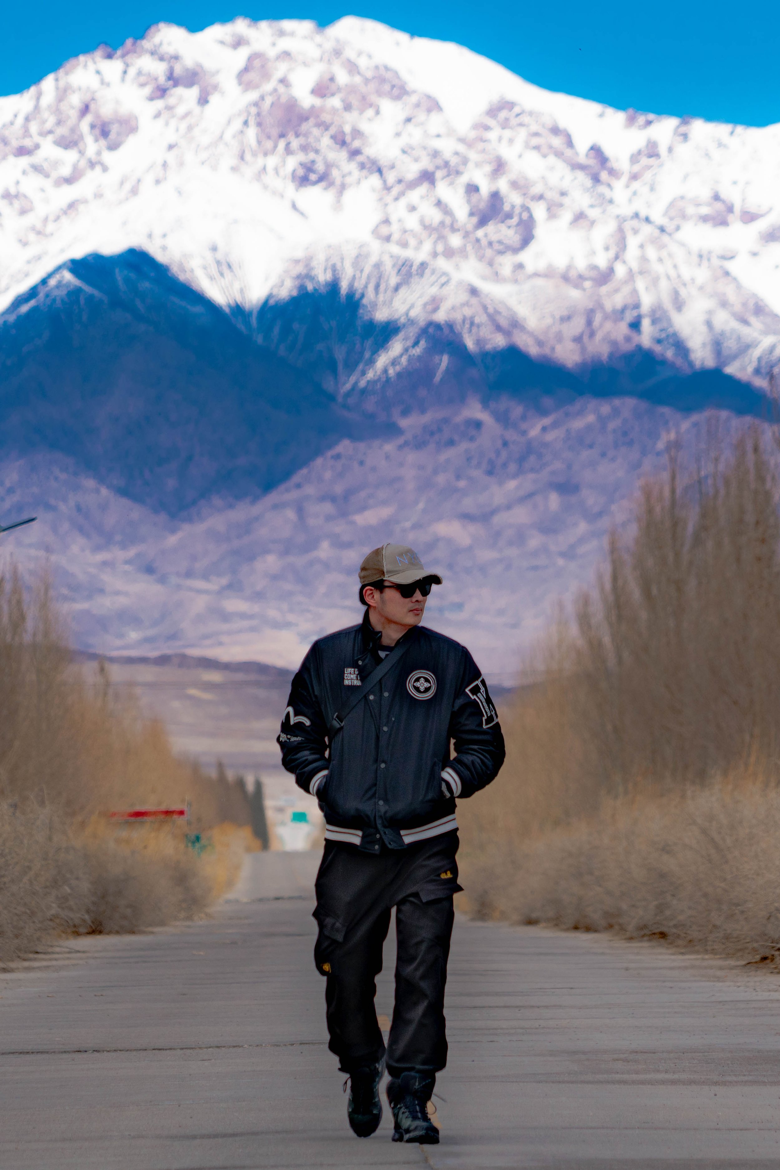 A man dressed in black with a jacket, pants, and sunglasses walking on a paved road with tall, dry grass on either side, with snow-capped mountains in the background under a clear blue sky.
