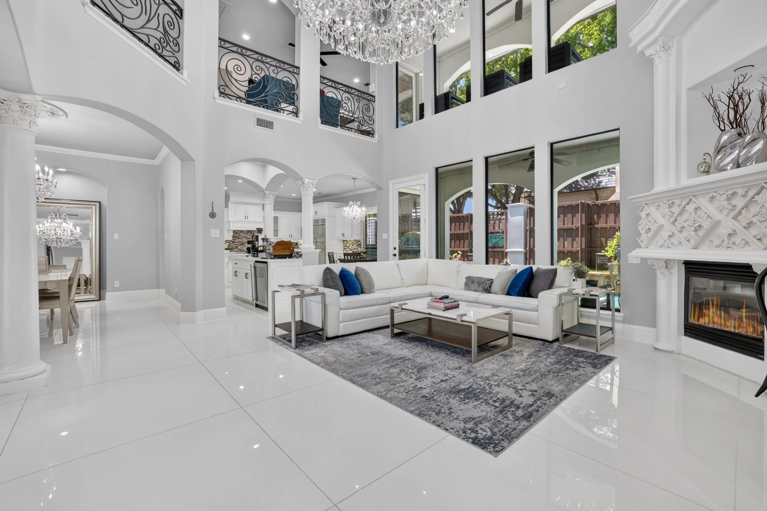 Bright, spacious living room with white walls, ceiling, and glossy tile floors. Features a large white sectional sofa with colorful pillows, a glass coffee table, and a decorative rug. There are large windows and sliding doors letting in natural light, with a view of a backyard. A grand crystal chandelier hangs from the high ceiling, and there is an ornate white fireplace with decorative molding. The open floor plan extends into a kitchen and dining area, with elegant chandeliers, white cabinetry, and stone backsplash.
