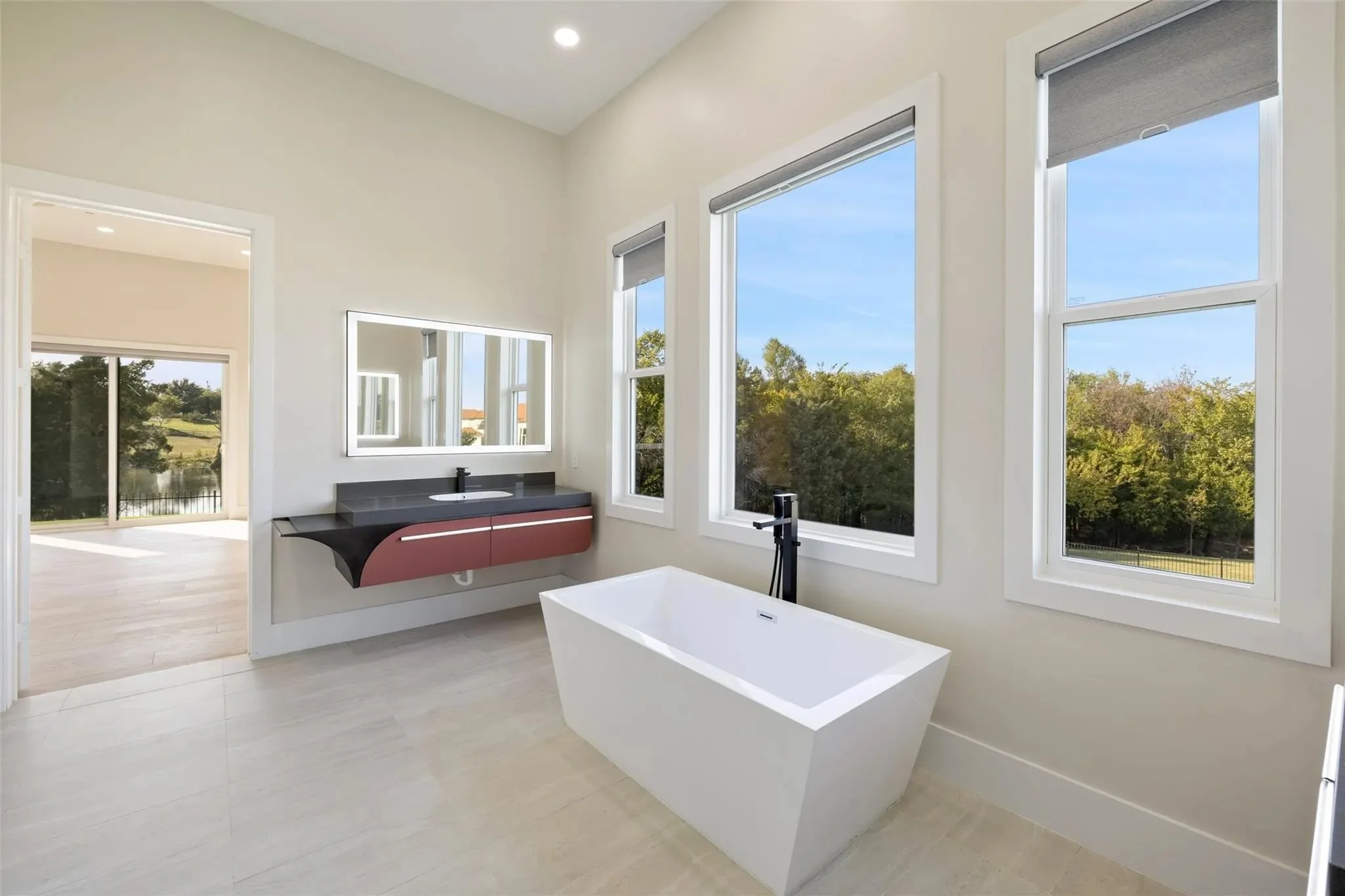Modern bathroom with a freestanding white bathtub, large windows showing trees outside, a floating vanity with a sink and mirror, and a view into an adjoining room with a sliding glass door.