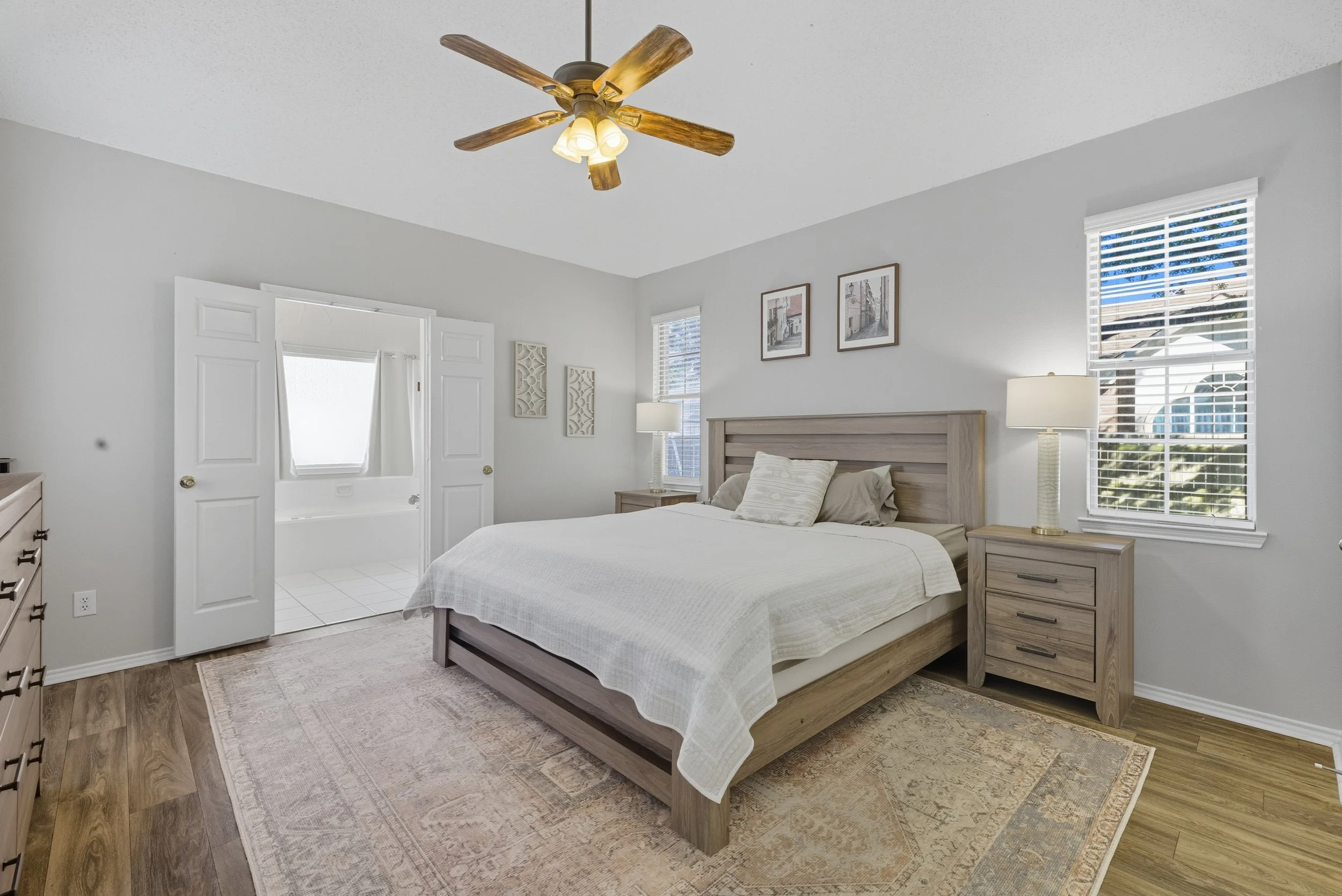 A bedroom with a wooden bed frame, two nightstands with lamps, a ceiling fan, and windows with blinds, connected to a bathroom with a bathtub.
