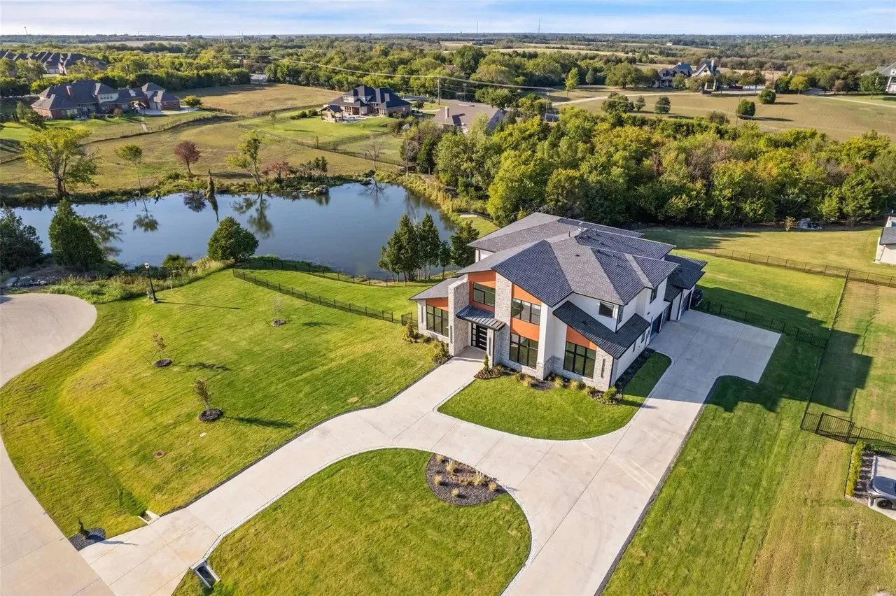Aerial view of a modern house with a large driveway and landscaped lawn, situated near a pond, with a rural landscape in the background.