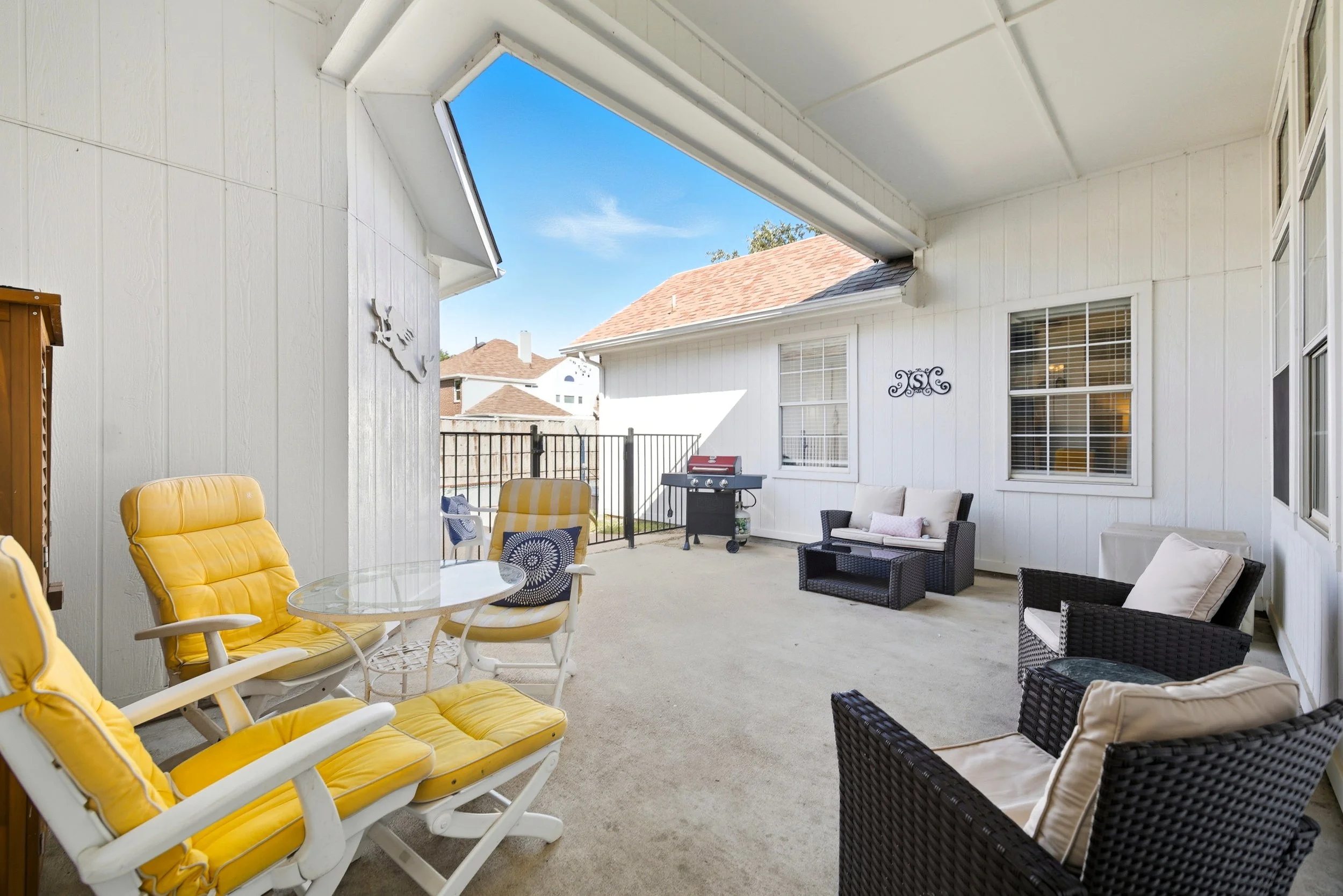 Covered outdoor patio with yellow and black wicker chairs, a glass-top table, a small black sofa, a black wicker armchair, a barbecue grill, and decorative initials "JSC" on the wall. The patio has a concrete floor and white siding walls, with the sk