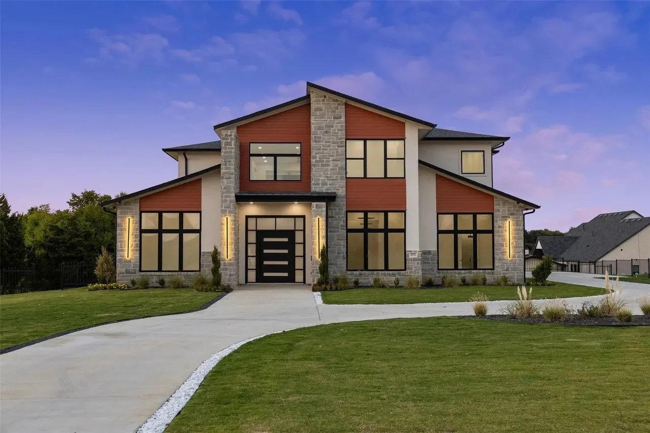 Modern two-story house with a stone and wood exterior, large windows, front yard with green grass and plants, and a curved concrete driveway at dusk.