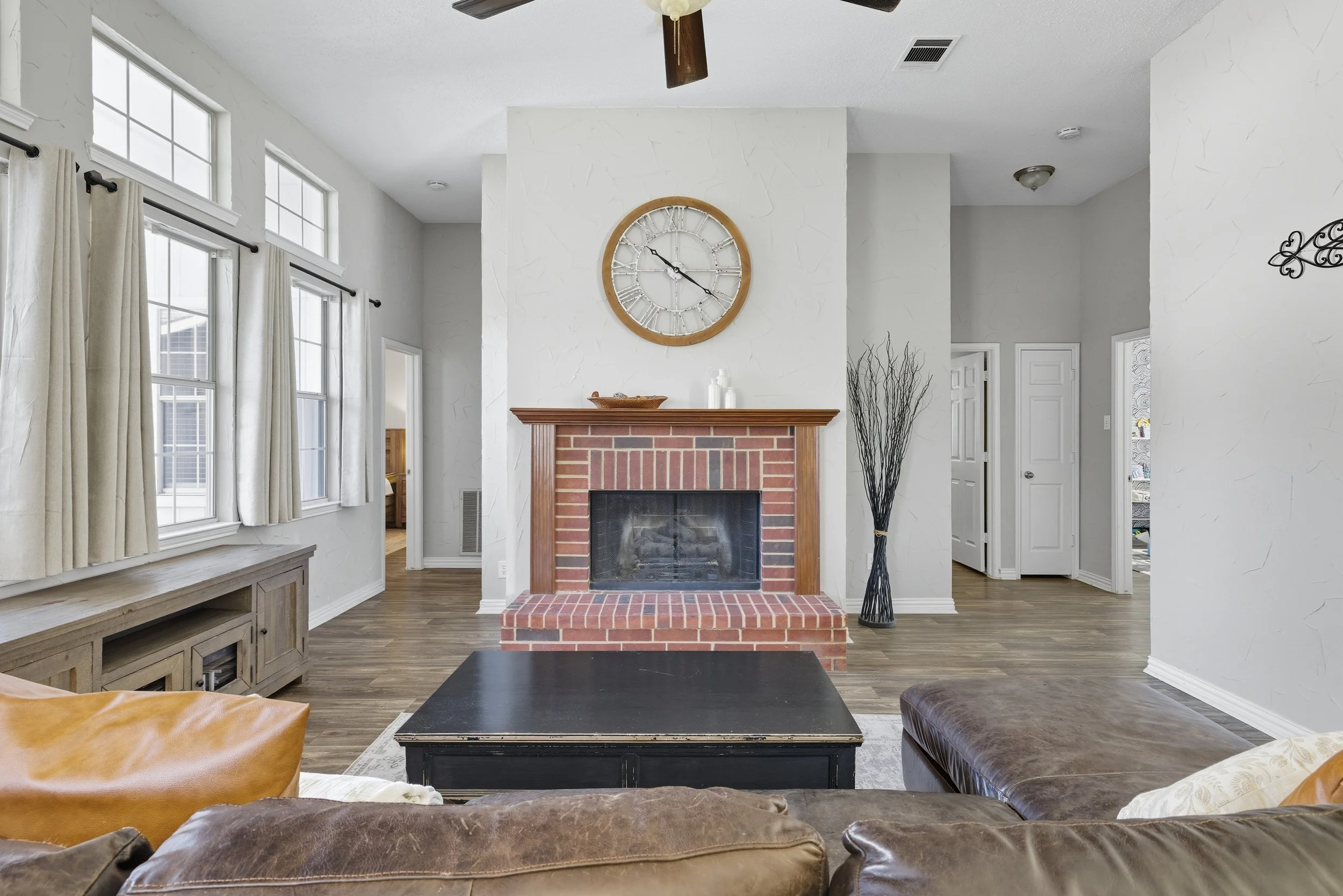 Living room with a brick fireplace, large wall clock, and dark furniture, bright windows with curtains, hardwood floors.