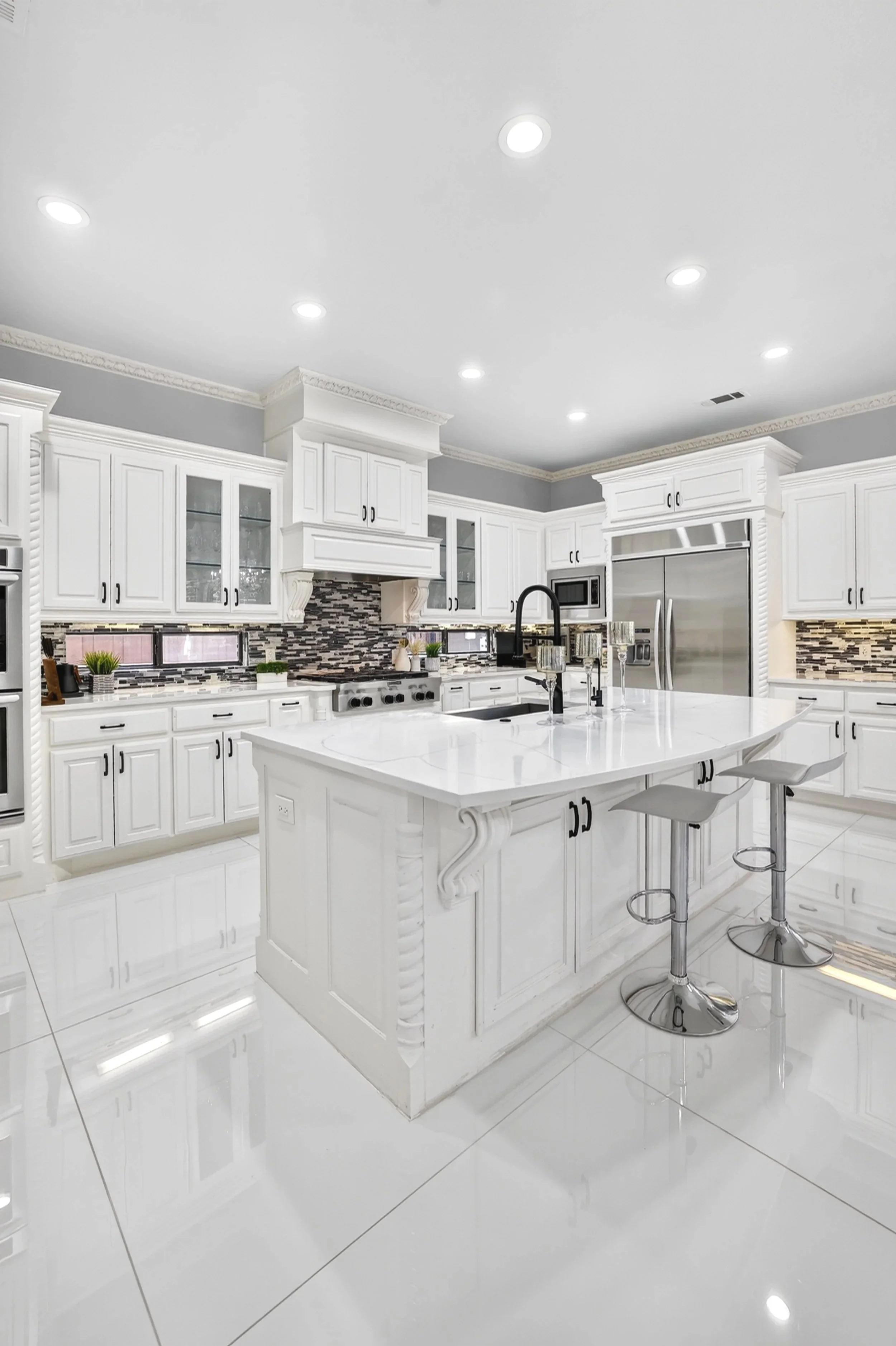 A bright modern kitchen with white cabinets, a large white island with a black sink, and a patterned backsplash. Two bar stools are at the island, and stainless steel appliances are visible.