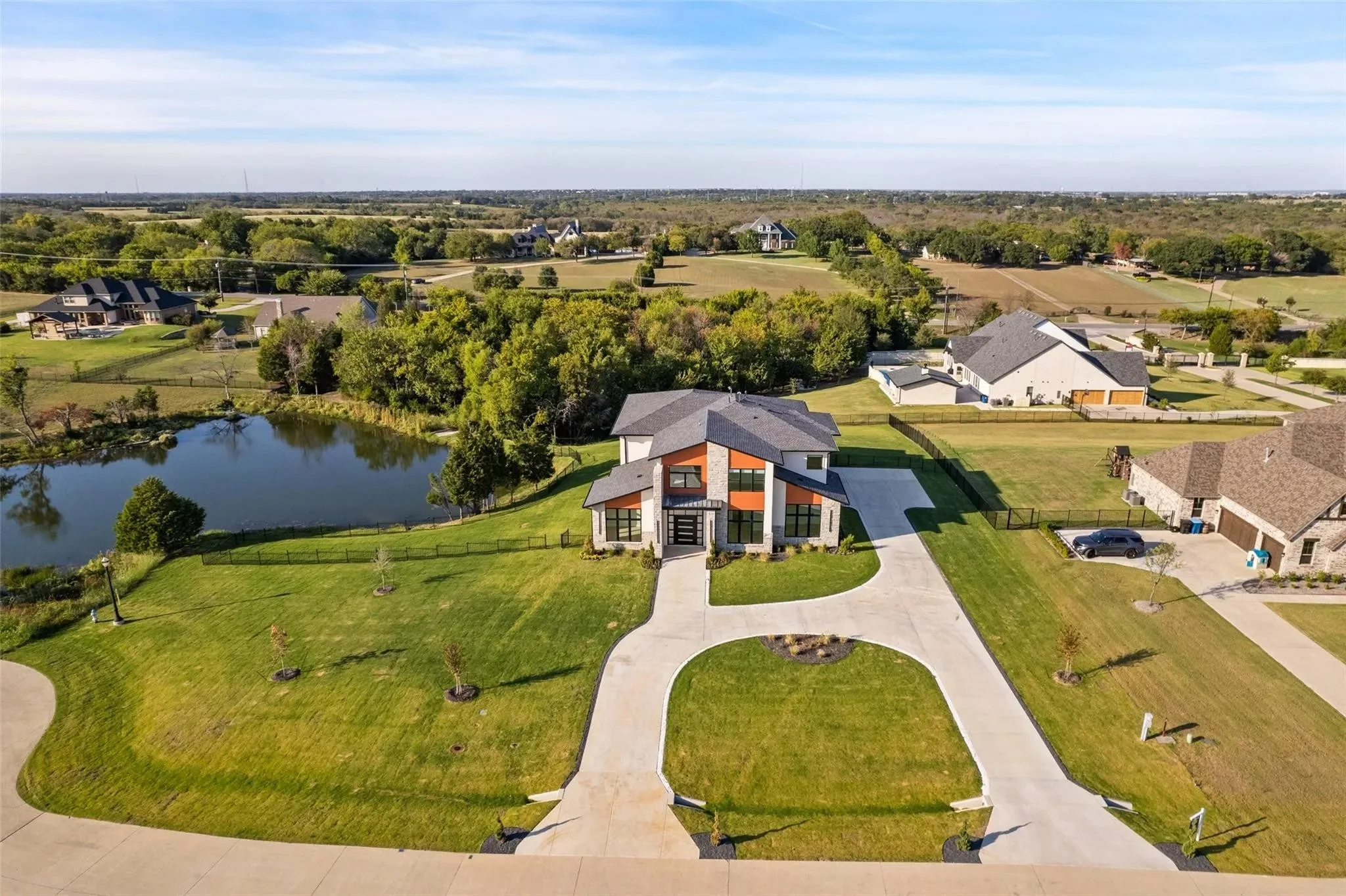 Aerial view of a modern house with a spacious driveway, green lawn, and a pond on the left, in a suburban neighborhood with other houses, trees, and open fields in the background.