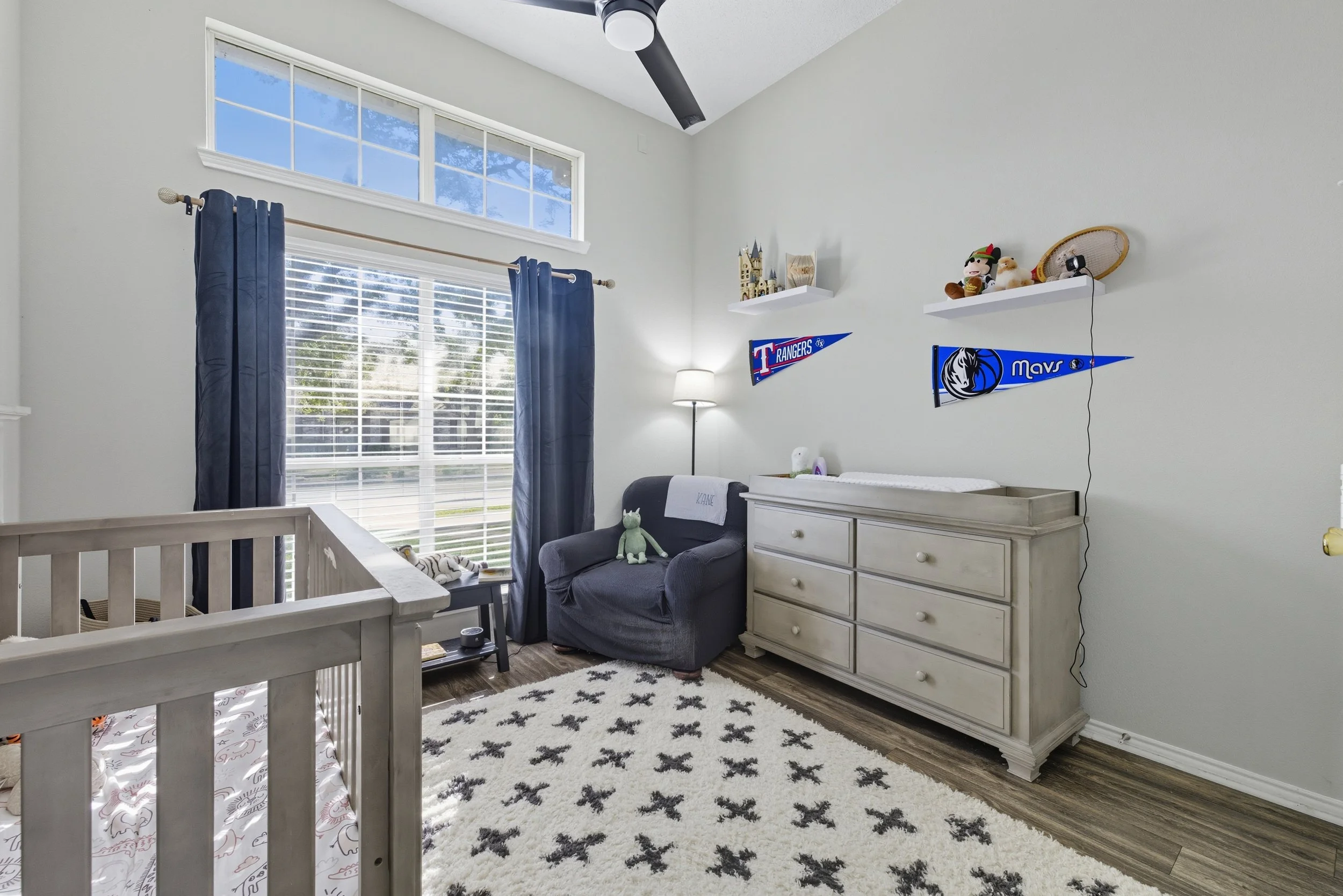 A baby nursery with a blue and white color scheme. Contains a crib, a chair with plush toys, a dresser, and sports pennants on the wall. The room has a large window with blinds, dark curtains, and a ceiling fan.