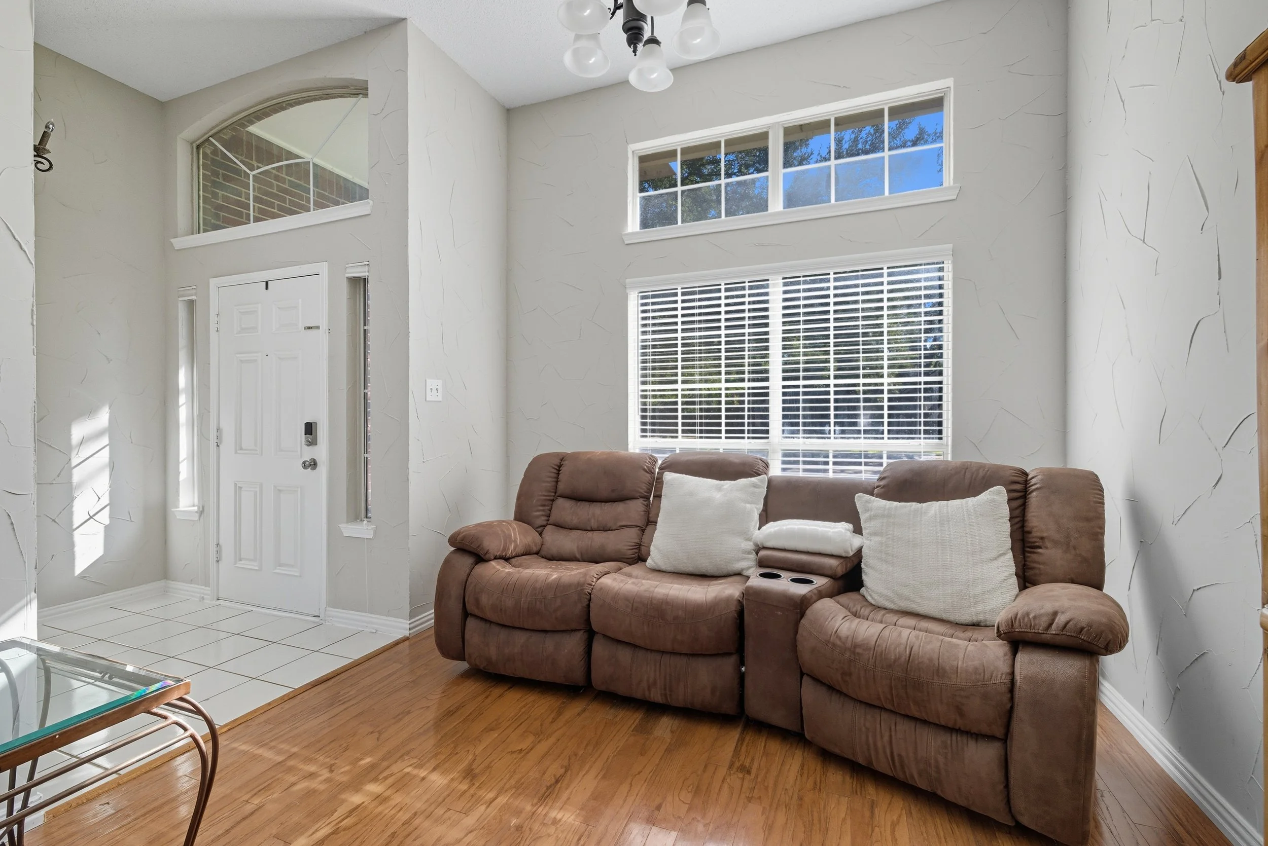 Living room with a brown recliner sofa, white pillows, hardwood floor, large window with white blinds, front door area, and half-moon window above the door.