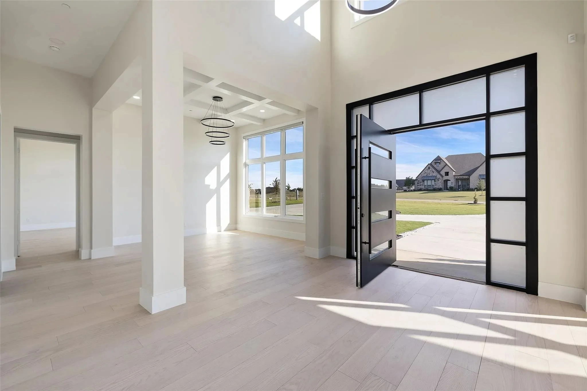 Empty modern living room with white walls, large front door with black frame, and big window showing a suburban neighborhood, bright sunlight.