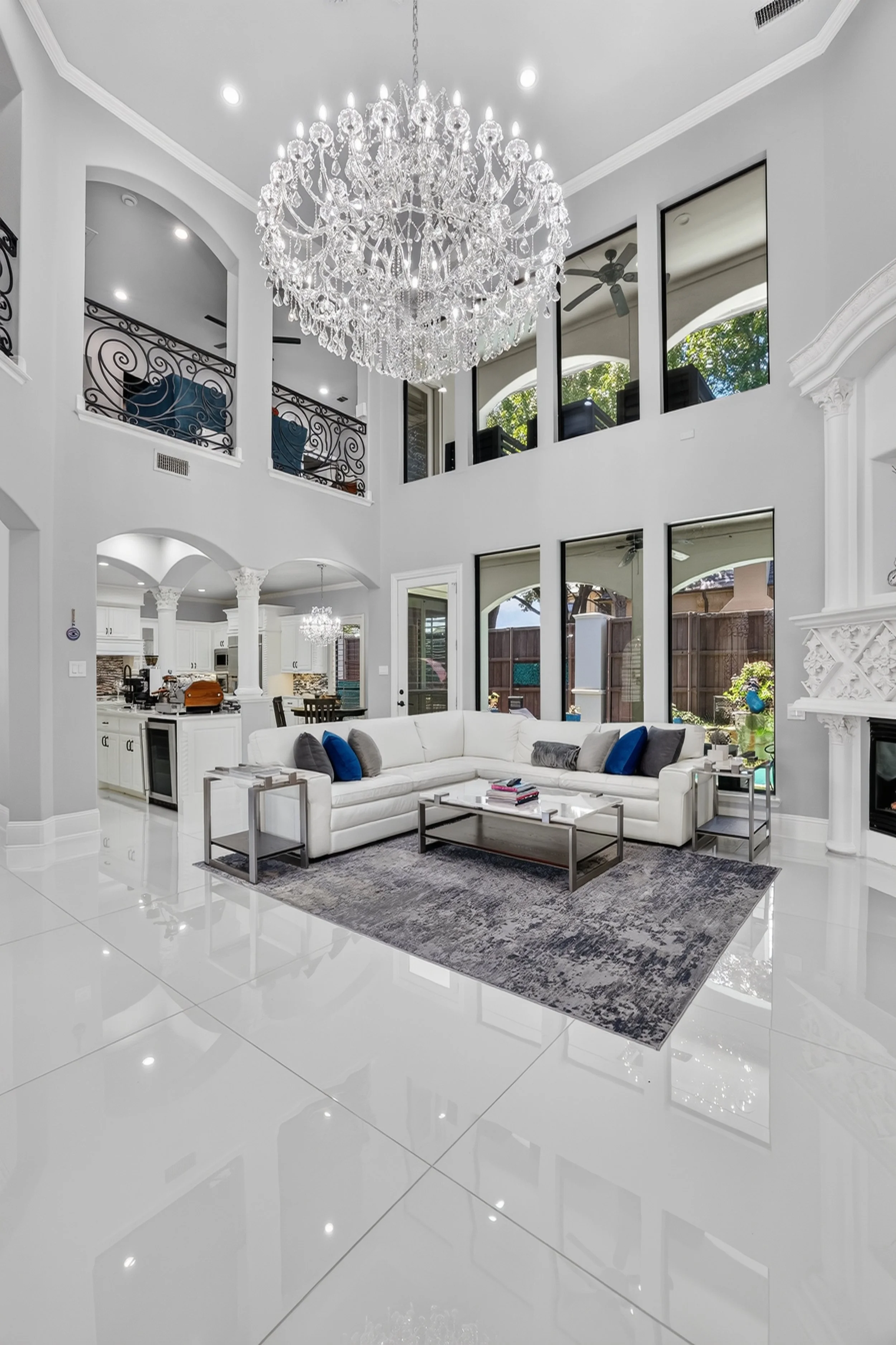 Interior of a modern living room with white walls, a large chandelier, a white sectional sofa with blue and gray pillows, a gray and white rug, and large windows providing natural light.