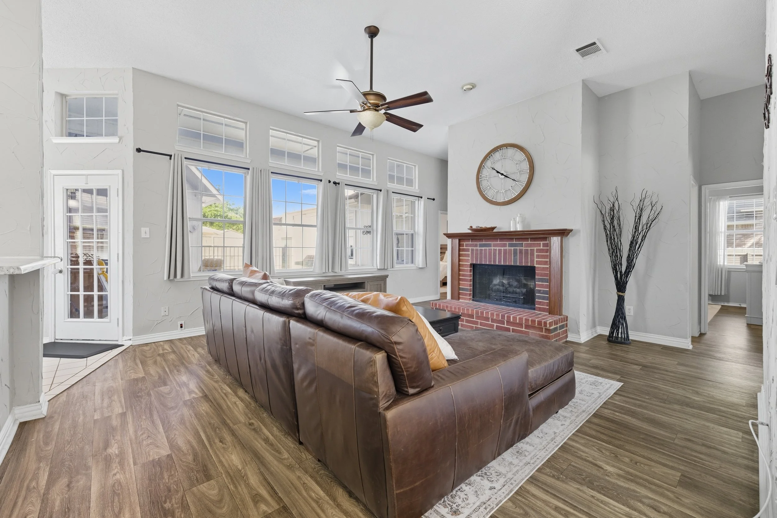 Living room with a brown leather sectional sofa, a brick fireplace, a round wall clock, and large windows with white curtains, wood flooring, and a ceiling fan.