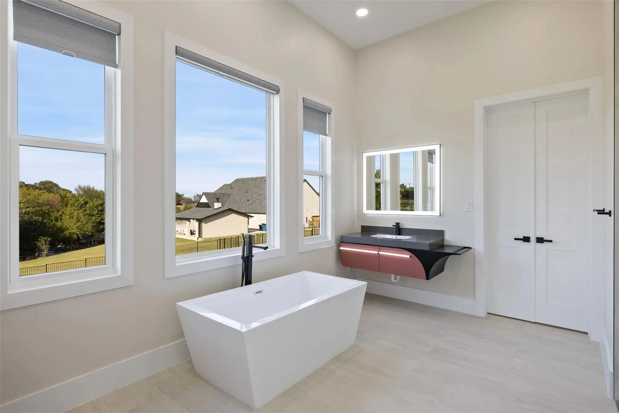 Modern bathroom with a white freestanding bathtub, large windows showing a backyard, a wall-mounted sink with a mirror above, and white double closet doors.