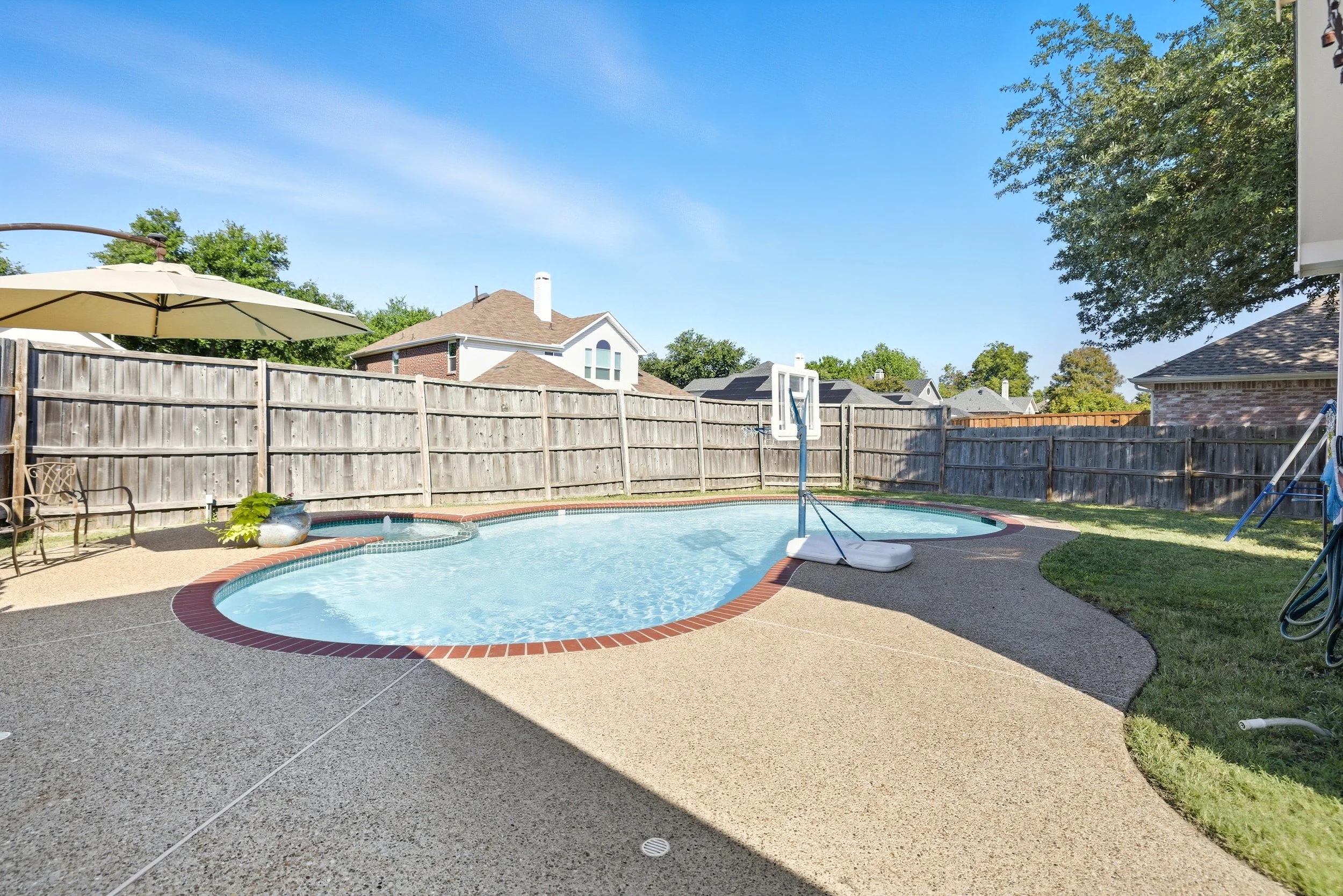 Residential backyard with a kidney-shaped swimming pool, patio furniture, a large umbrella, a basketball hoop, and a wooden fence, under a clear blue sky.
